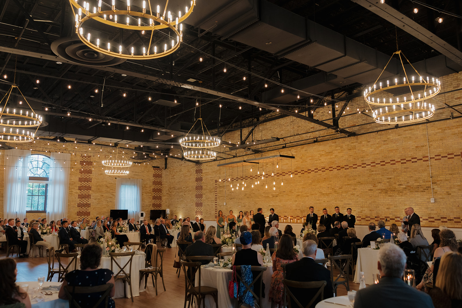 A spacious wedding reception hall adorned with elegant chandeliers, filled with guests celebrating together.