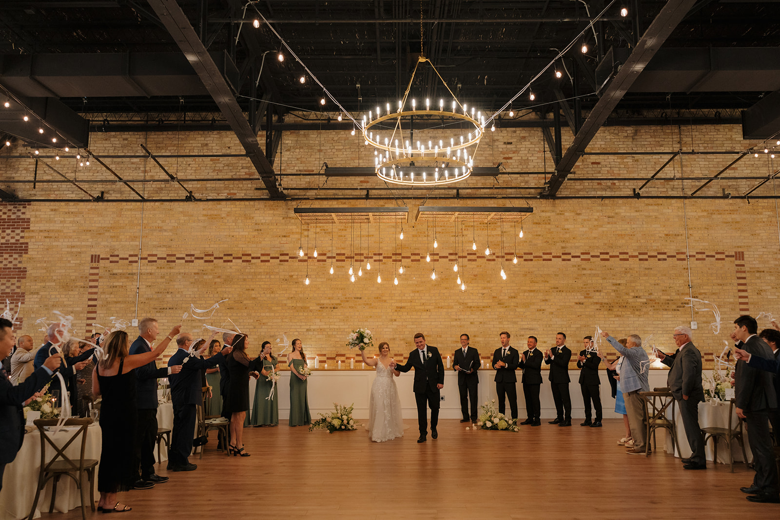 Guests gather for a wedding ceremony in a grand room, highlighted by stunning chandeliers. Bride and groom celebrate recent marriage by lifting hands in the air