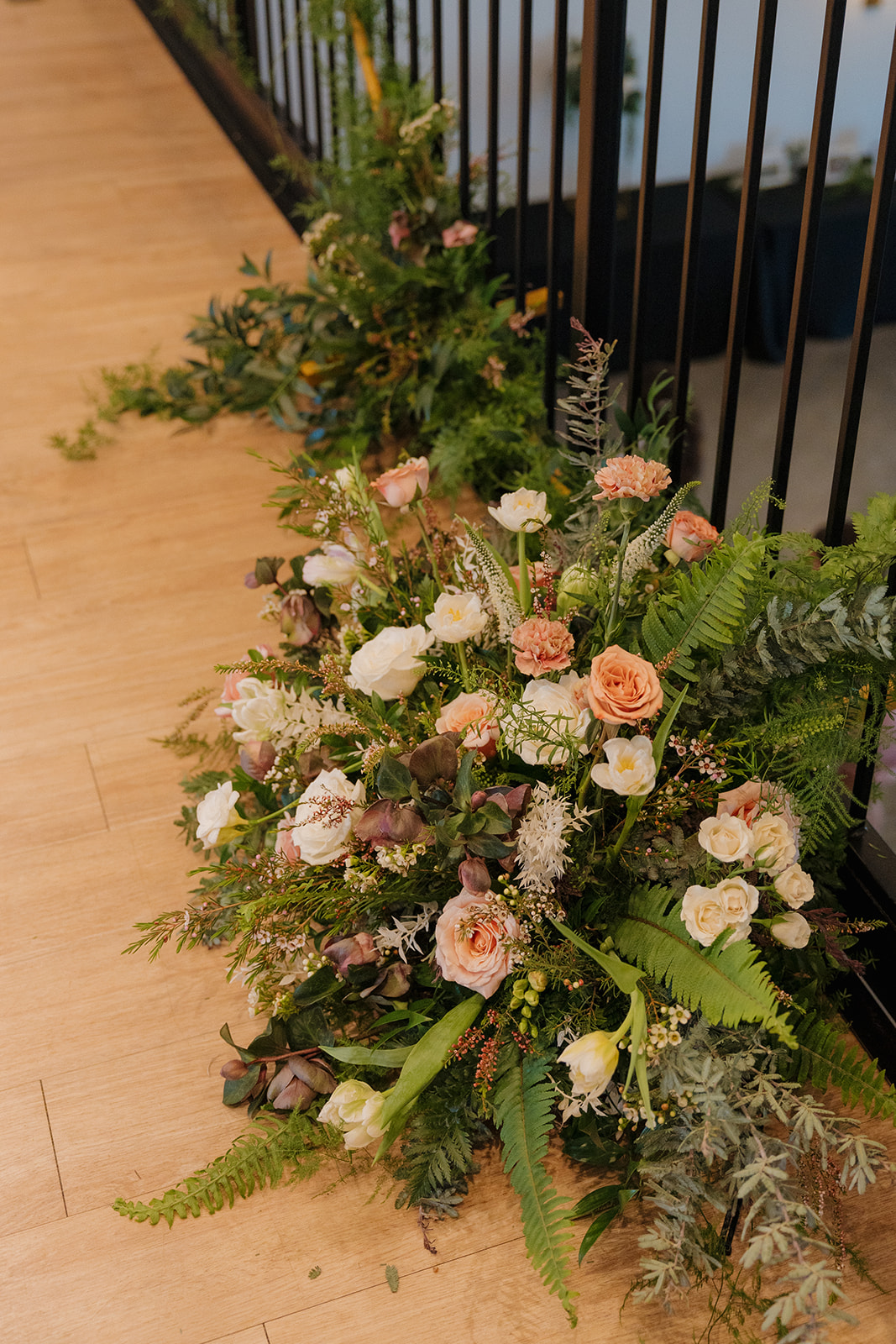 Lush ground arrangements of greenery, white roses, and blush-toned florals line the ceremony aisle against modern black railing.