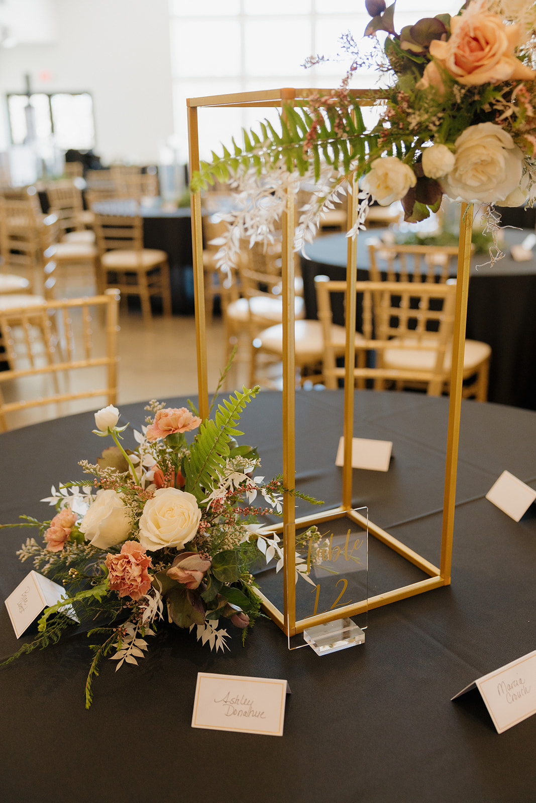Elegant floral arrangement in a geometric gold frame, surrounded by blush roses and name cards on a black tablecloth.