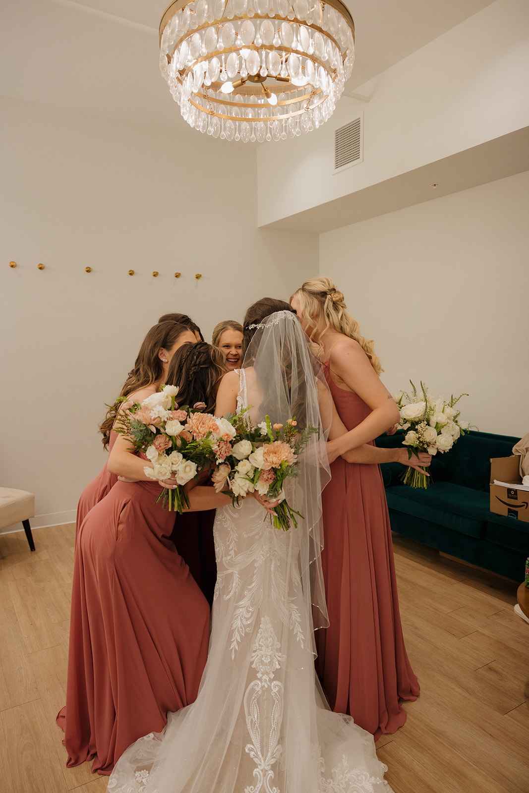 Bride and bridesmaids share a tight pre-ceremony group hug, bouquets in hand beneath a sparkling chandelier—madison wedding photographer captures the love and anticipation.