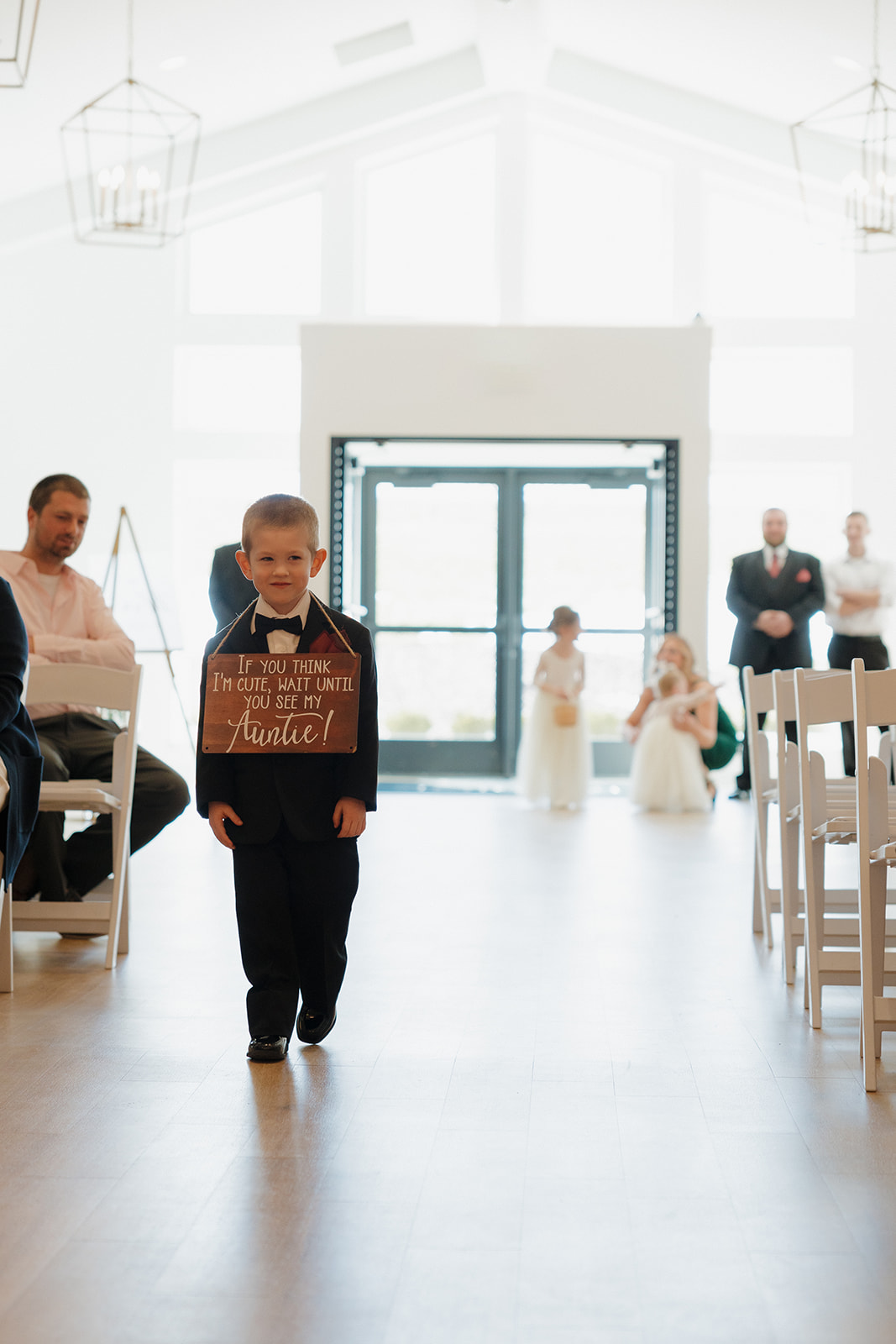 Young ring bearer walks down the aisle holding a wooden sign, with flower girls and guests in the background—madison wedding photographer documents every sweet detail.