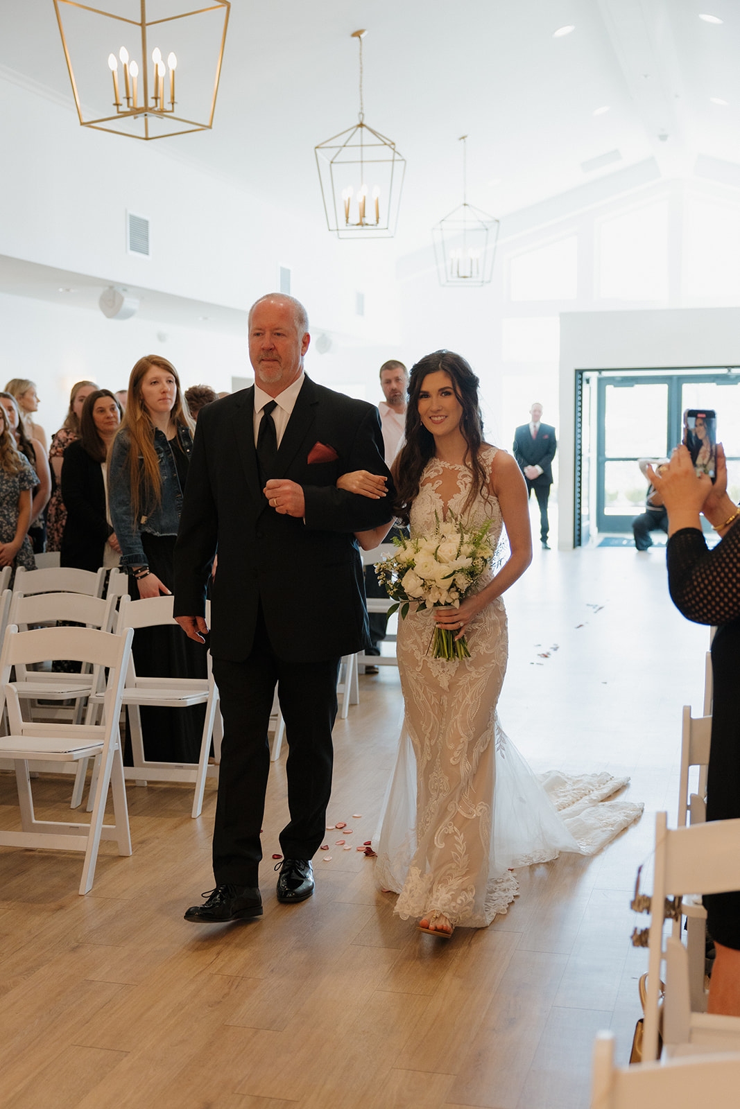 Bride walks arm-in-arm with her father down the aisle, holding a white bouquet and smiling as guests look on.