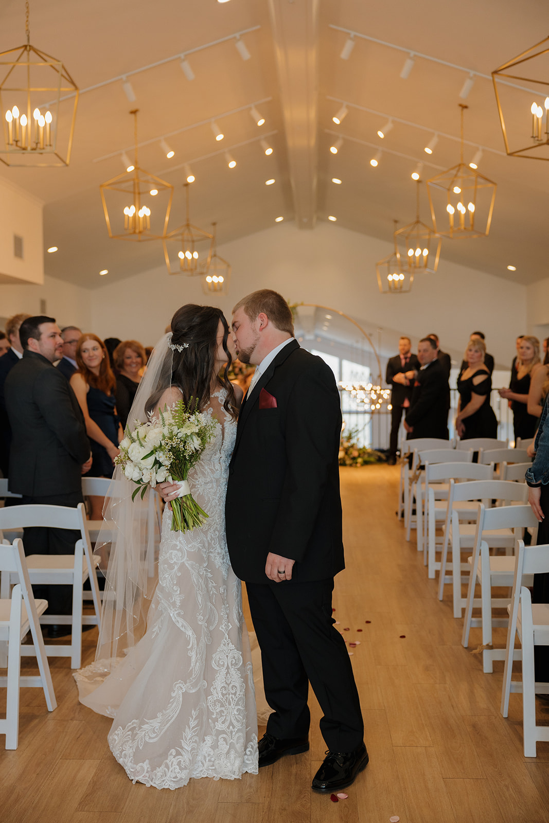 Bride and groom share a sweet kiss while walking back down the aisle in a modern white chapel, surrounded by guests and golden chandeliers.