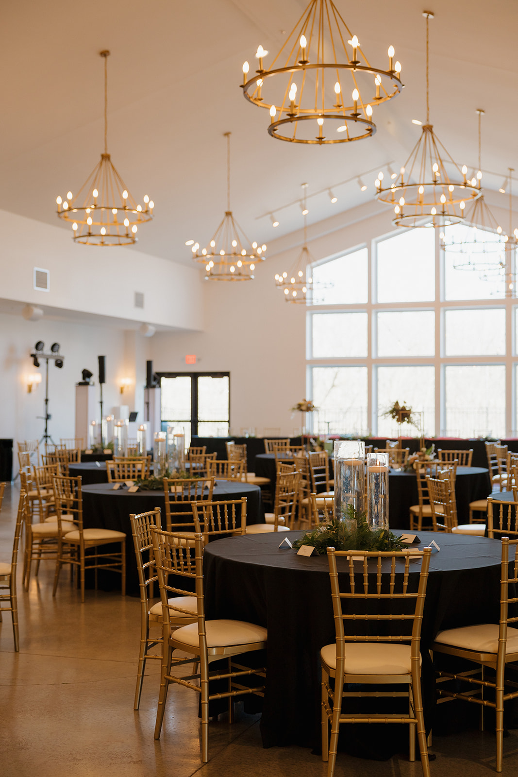 Reception hall set with black tablecloths, gold chiavari chairs, and floating candle centerpieces beneath grand chandeliers.