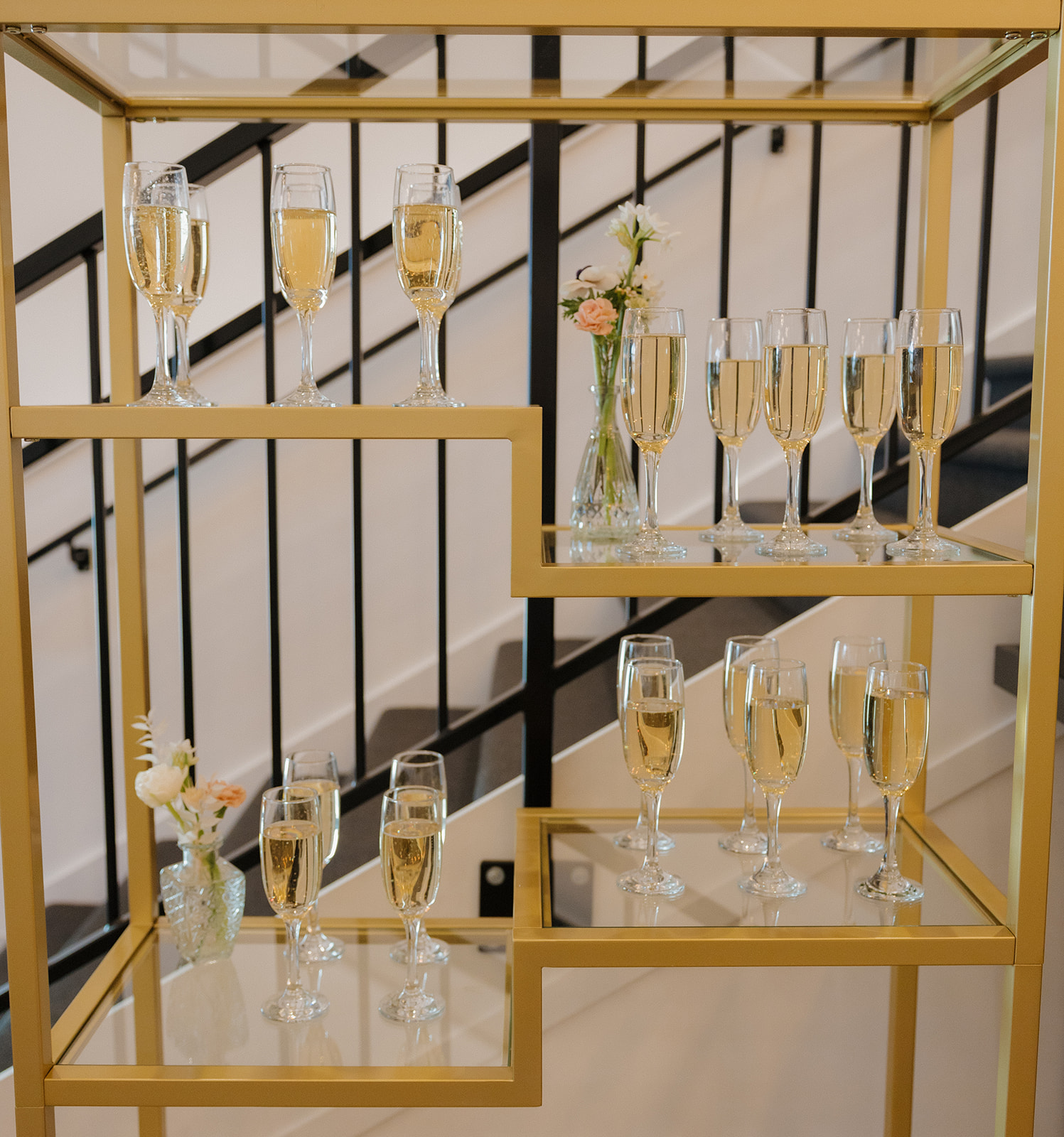 Rows of champagne flutes arranged on a modern gold display shelf, with soft florals and a chic black railing backdrop.