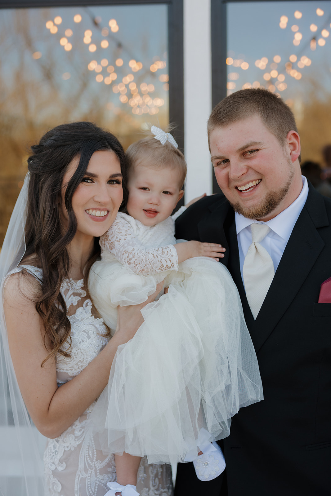 Bride and groom pose outside with their smiling daughter in her white dress, lit by soft string lights and sunshine—madison wedding photographer captures a sweet family moment.