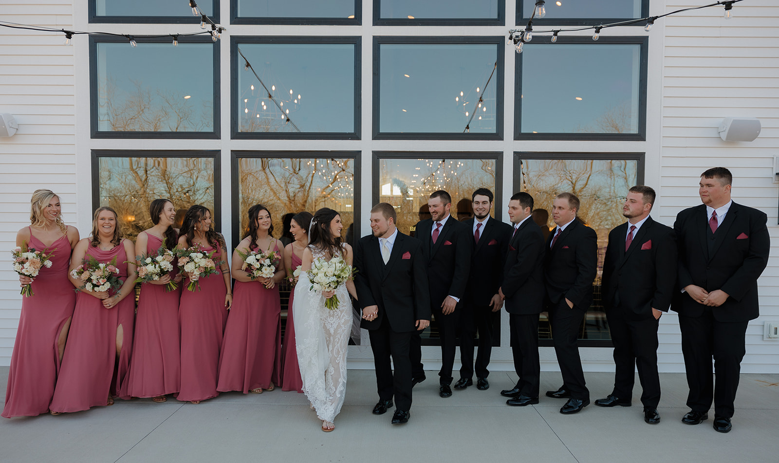 Bridal party lines up outside a modern white venue, with pink bridesmaid dresses and black suits framing the couple—madison wedding photographer documents the whole crew.