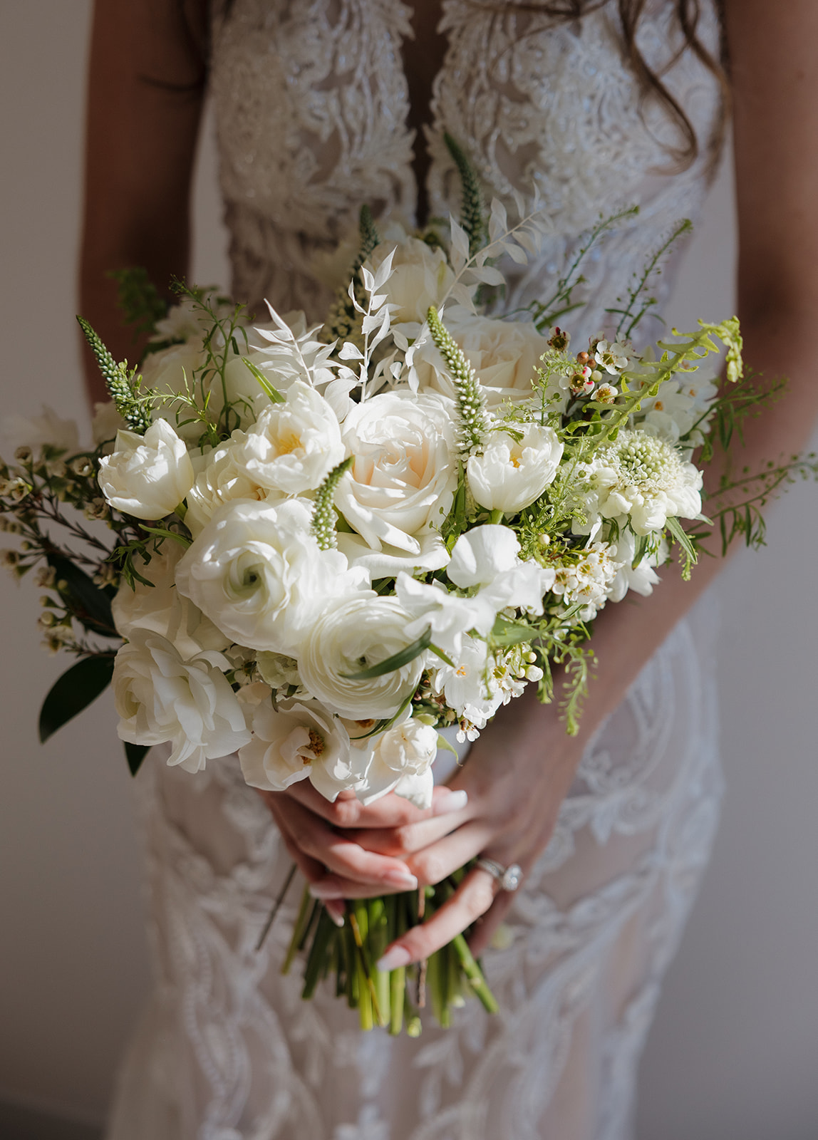 Bride holds a hand-tied bouquet of white ranunculus, garden roses, and greenery—soft shadows highlight every petal.