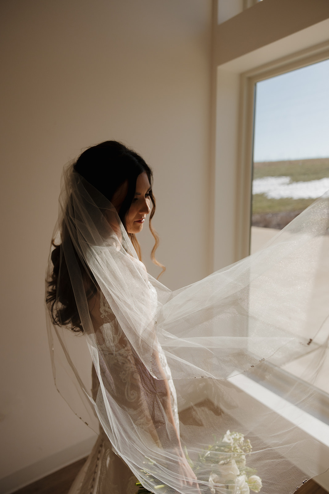 Bride stands peacefully by a window, soft light pouring in as her veil lifts mid-air—classic editorial bridal moment photographed by a madison wedding photographer.