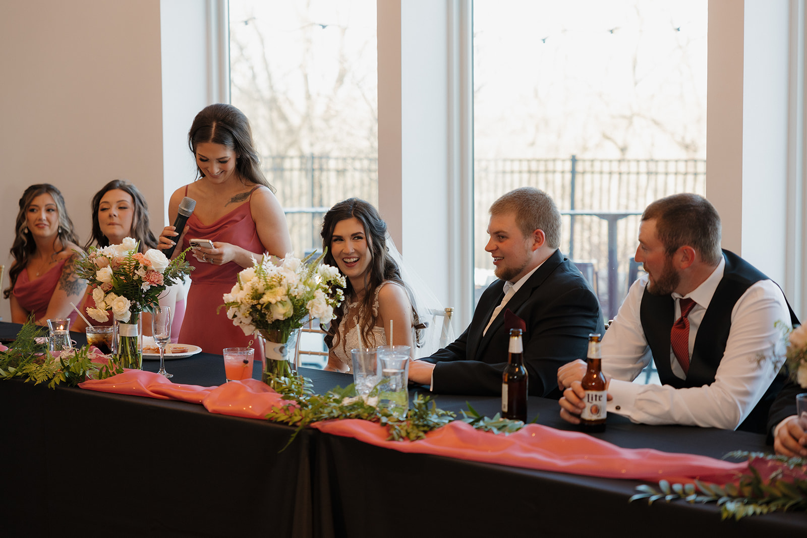 Maid of honor gives a toast while the bride and groom laugh at the head table, surrounded by florals and warm light.