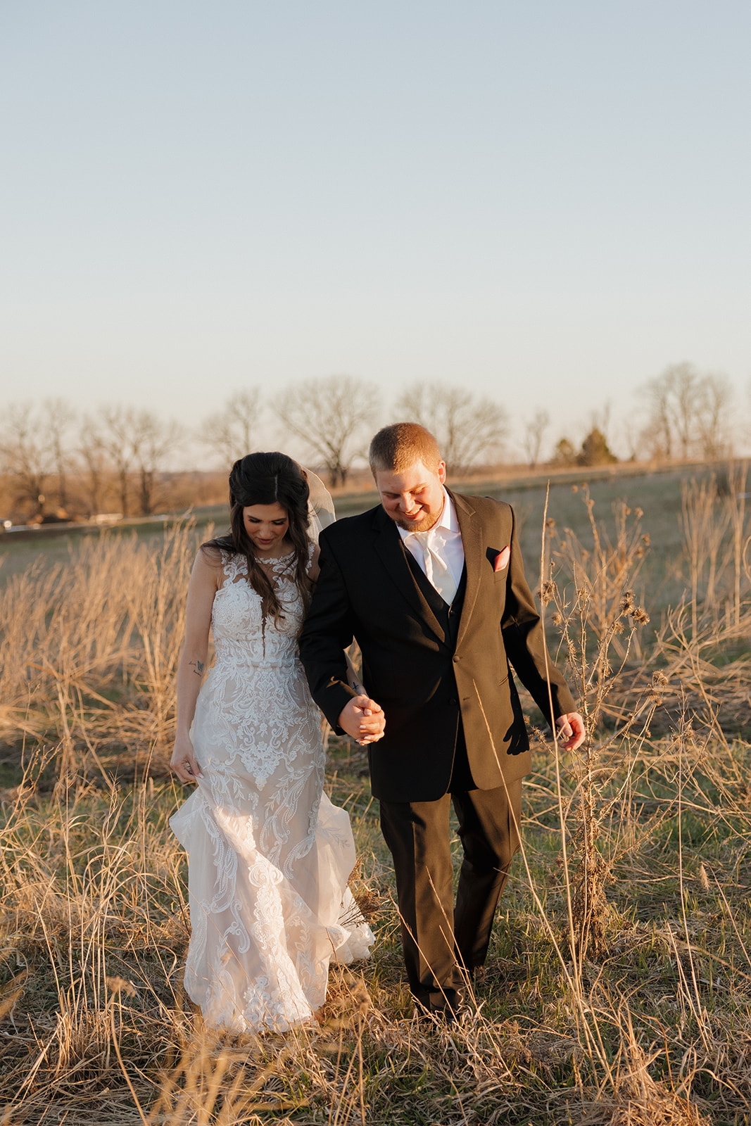 Newlyweds hold hands and laugh while strolling through dried grass at golden hour, dress hem catching the light.