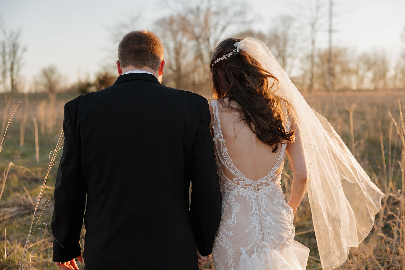 Bride and groom walk hand-in-hand through a rustic field, her veil catching the breeze as the sunlight glows behind them—captured by a madison wedding photographer.