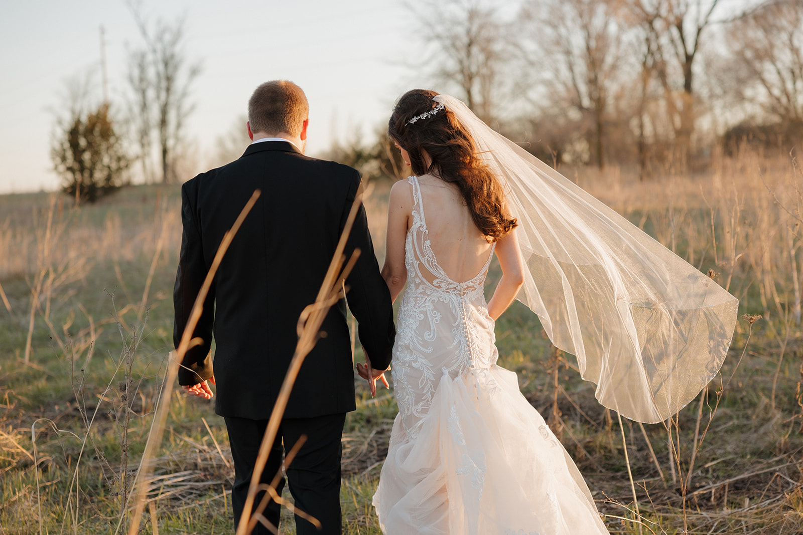 Bride and groom walk hand-in-hand through a golden field at sunset, her veil trailing behind—another timeless frame from a madison wedding photographer.