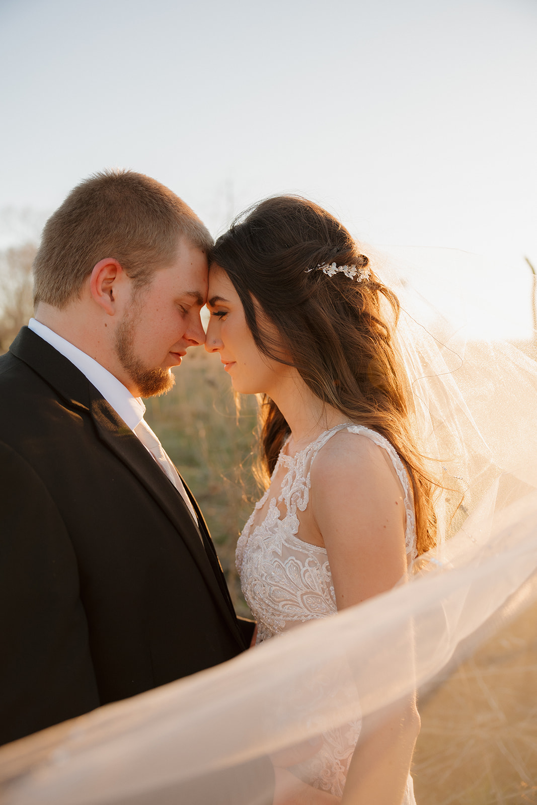 Bride and groom rest their foreheads together beneath her flowing veil, bathed in golden light during sunset portraits.