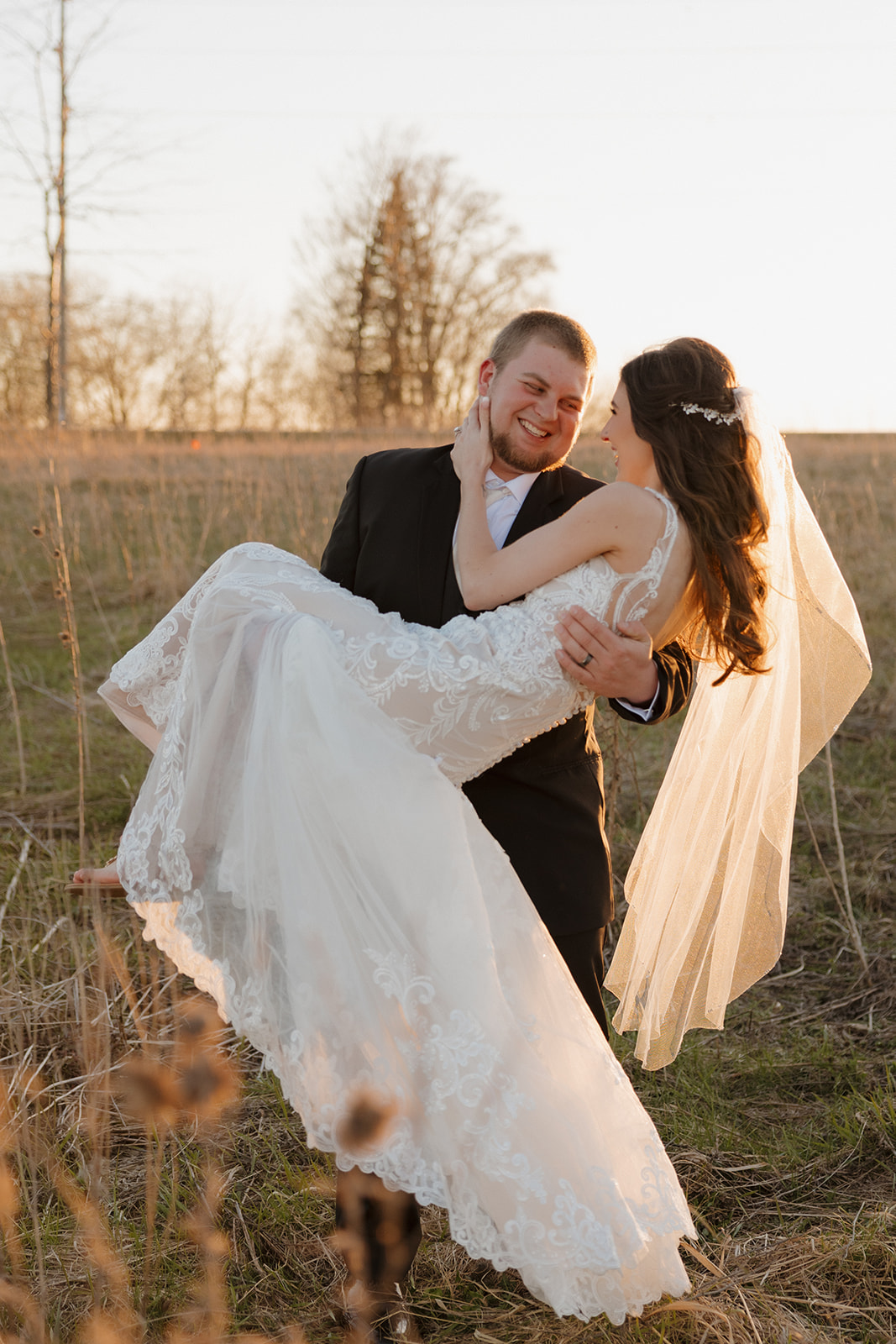 Groom lifts his bride in a golden field at sunset, both laughing as her lace gown flows—pure post-ceremony joy.