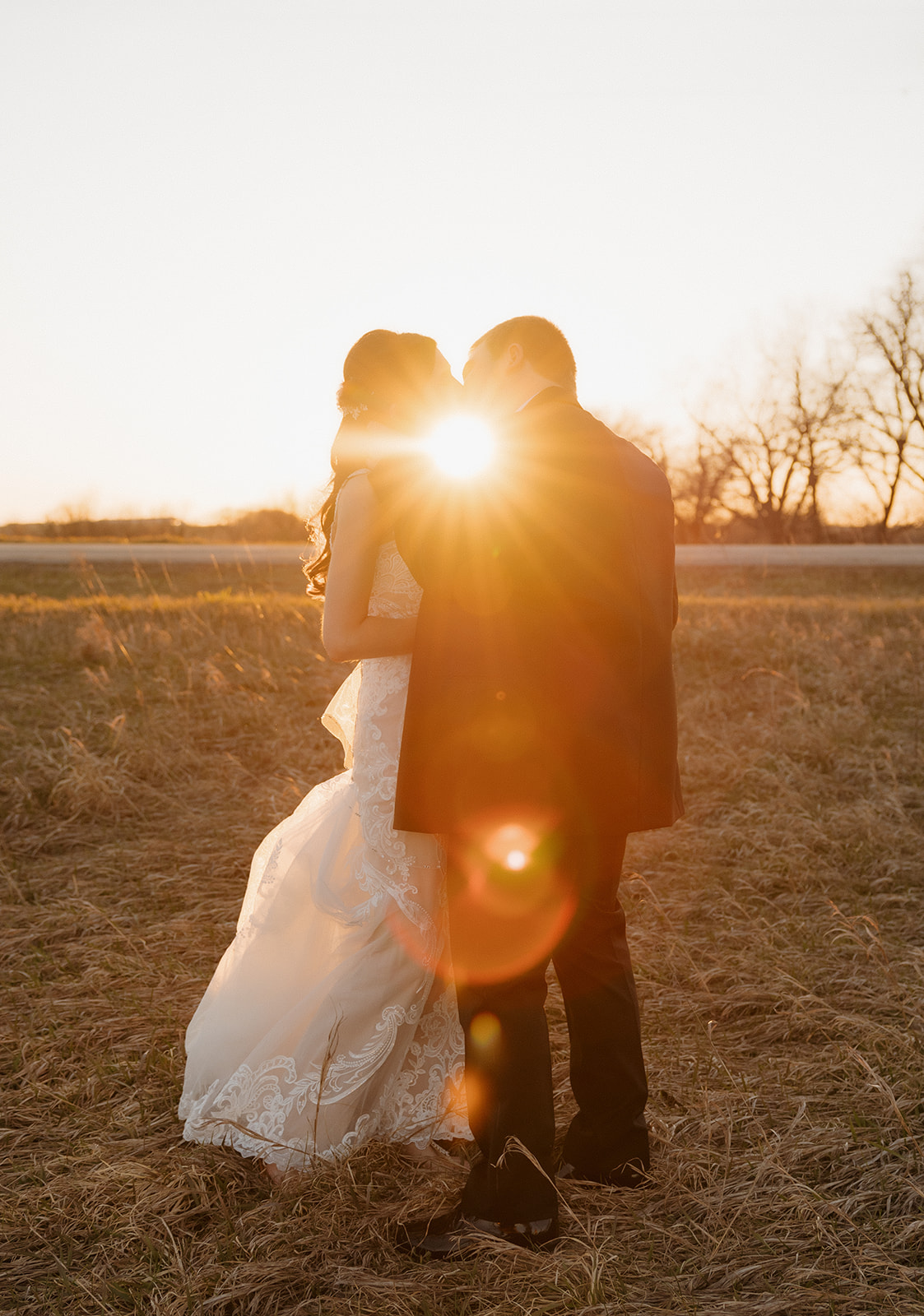 Bride and groom share a kiss in a golden field as the sun flares dramatically behind them—dreamy light meets real love.