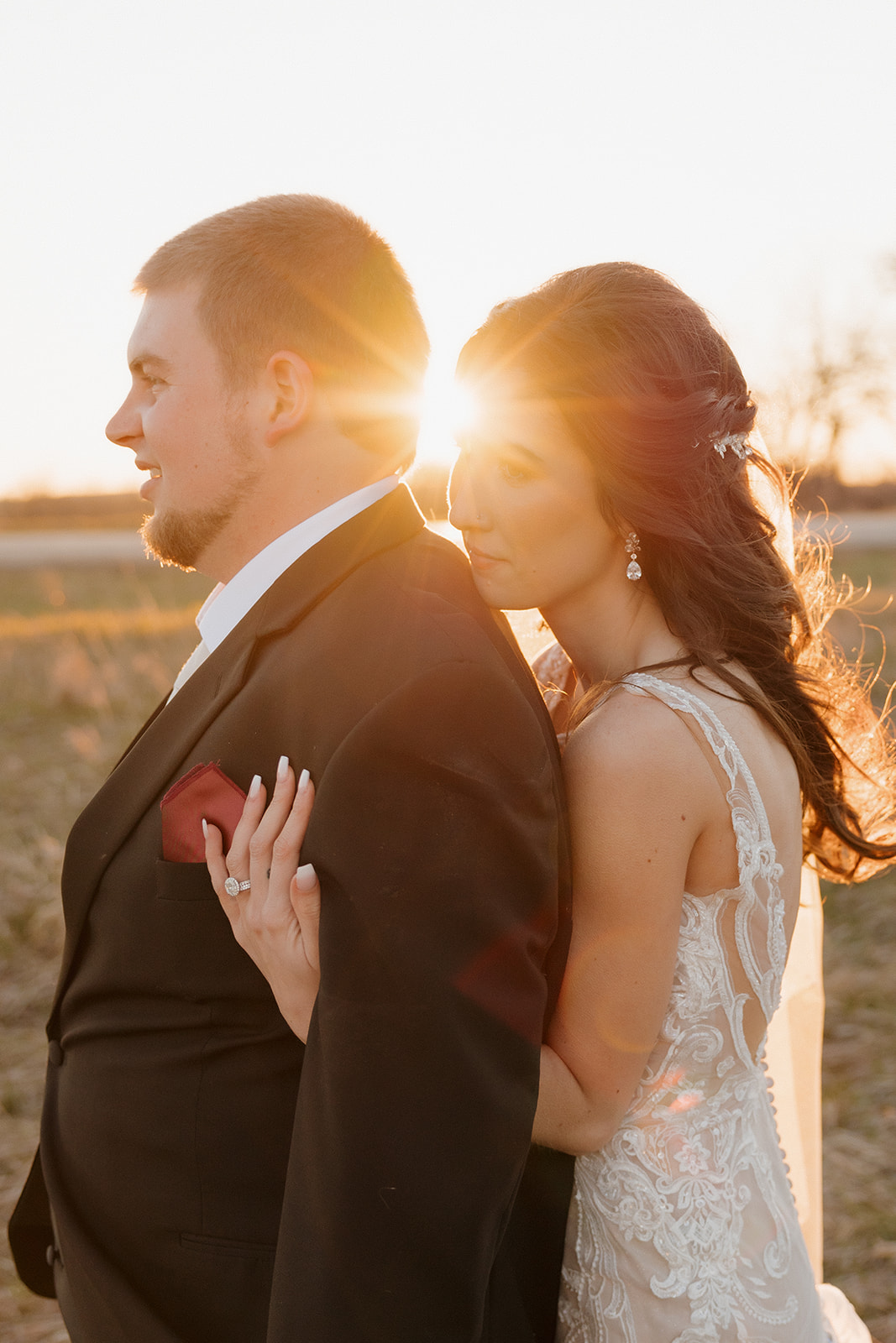 Bride hugs the groom from behind with the setting sun flaring between them—soft, golden romance in a quiet field.