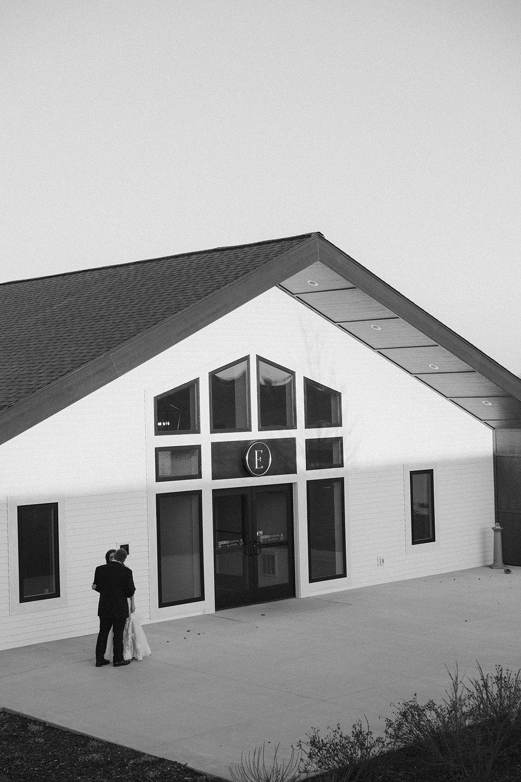 Black and white photo of the couple standing in front of a modern white chapel with triangular windows and clean lines.