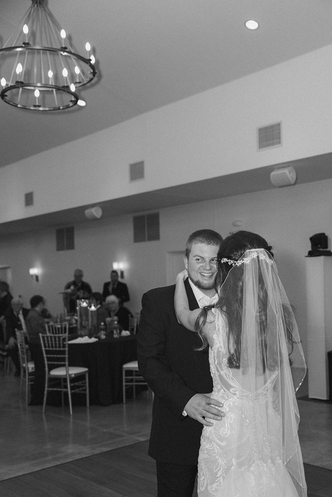 Black and white photo of the bride and groom mid-first dance, beaming at each other under glowing chandeliers.