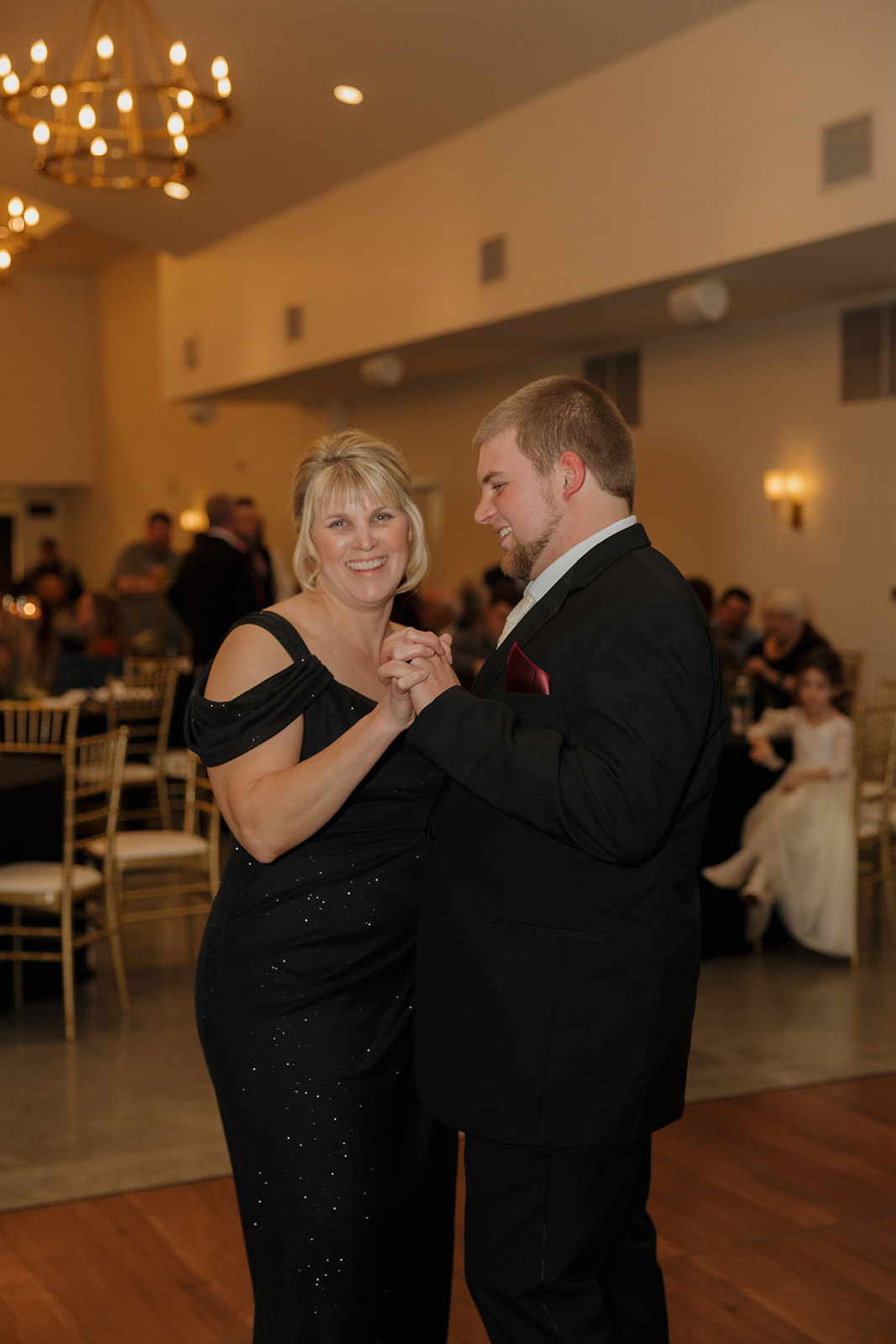 Groom shares a heartfelt mother-son dance surrounded by guests at a warmly lit reception.