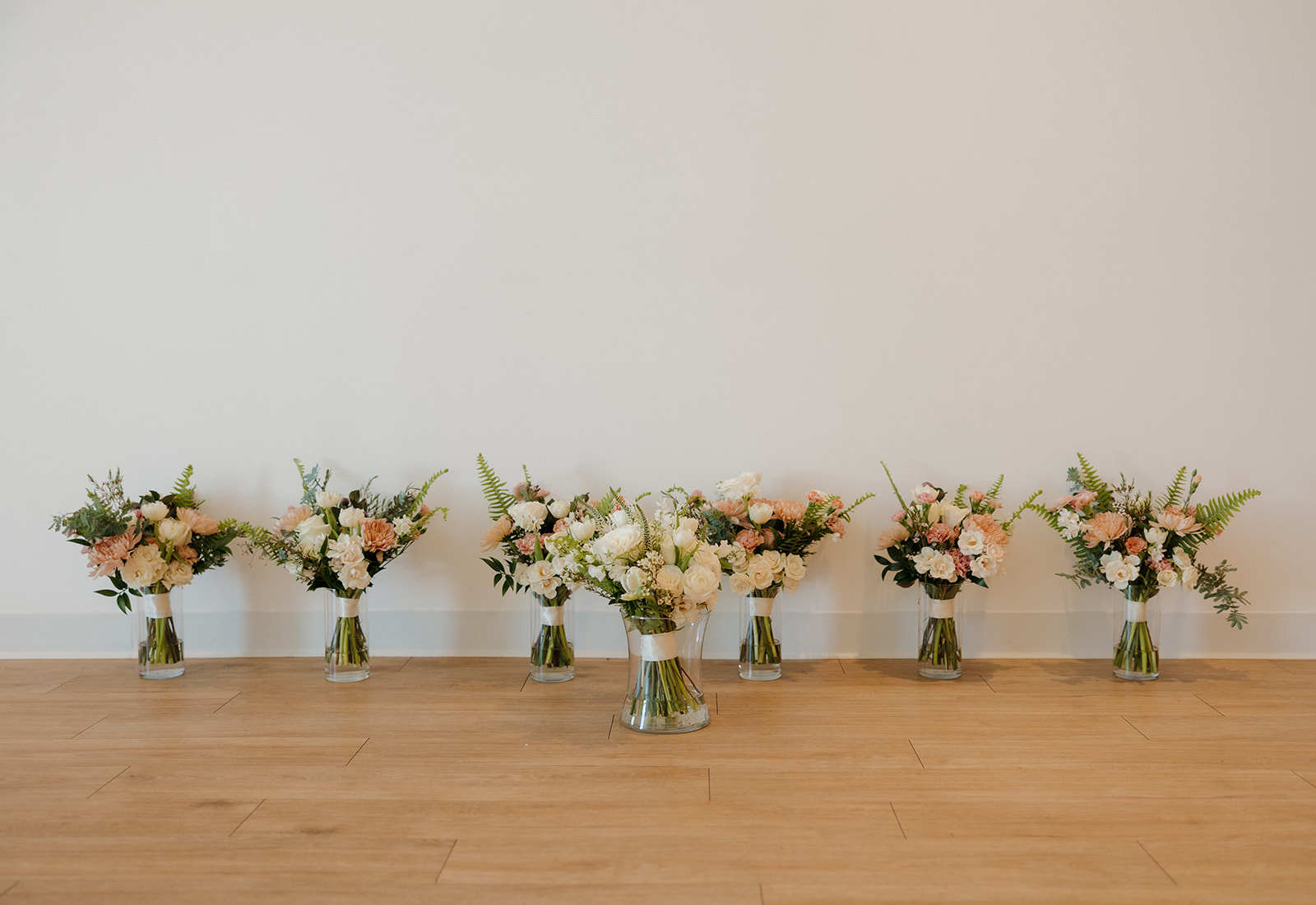 Seven lush bridal bouquets in glass vases lined up against a clean white wall, featuring soft blush, ivory, and greenery.