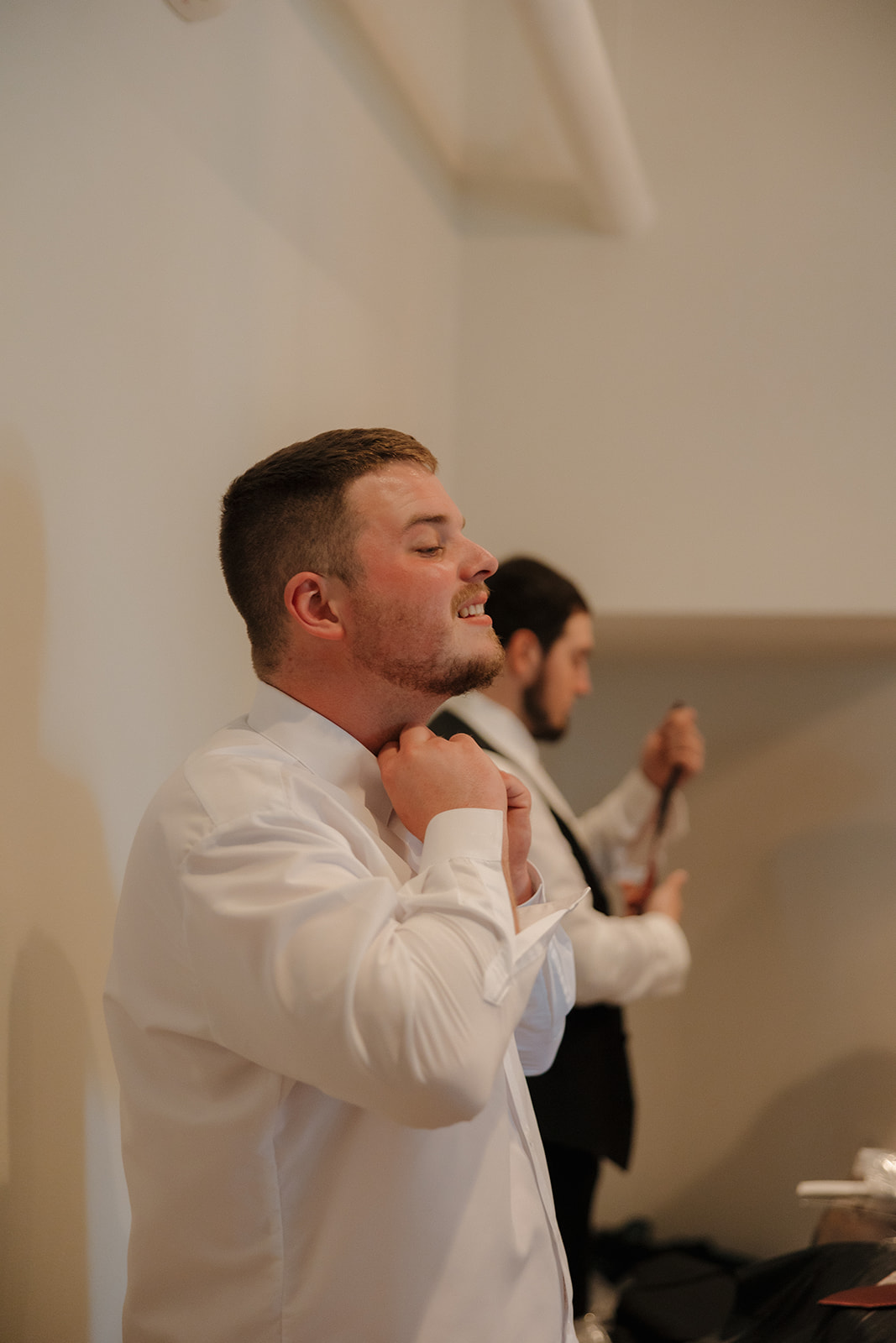 Groom adjusts his collar in a candid getting ready moment, with a groomsman fixing his tie in the background—classic prep vibes.