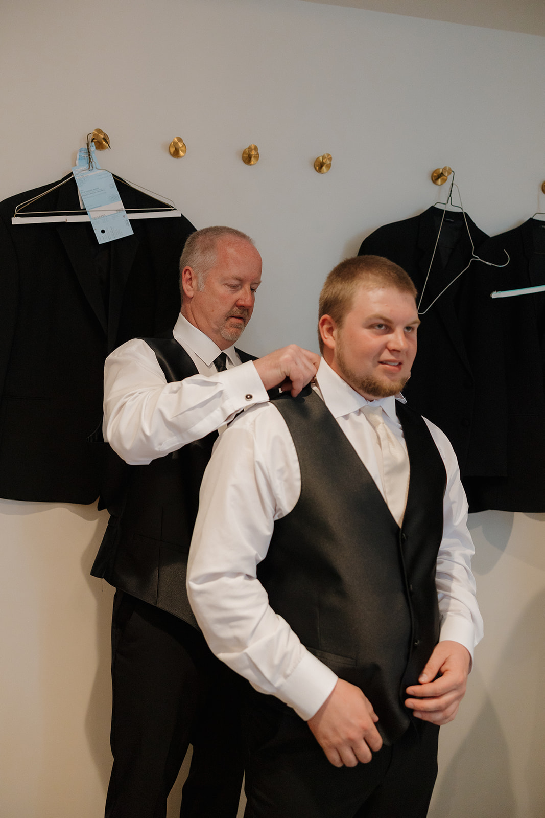 Father helps the groom into his vest before the ceremony, both dressed in crisp white shirts and black formalwear.
