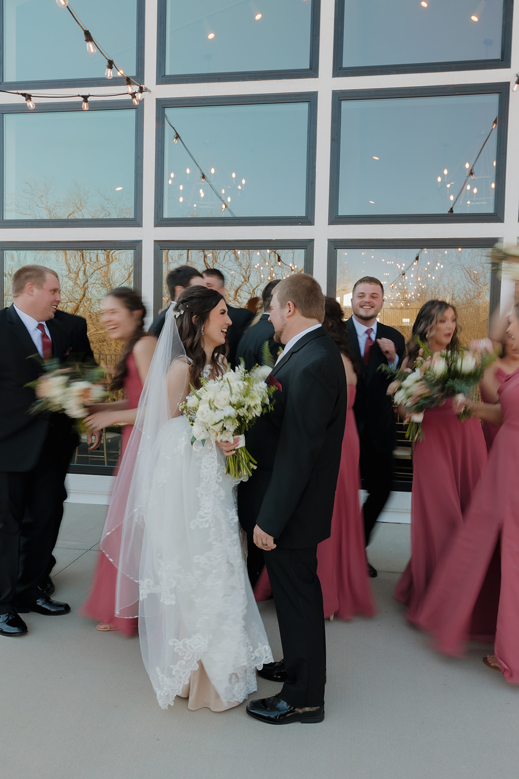 Bride and groom share a private smile while the bridal party swirls around them in motion—pure joy and movement captured outside their modern venue.