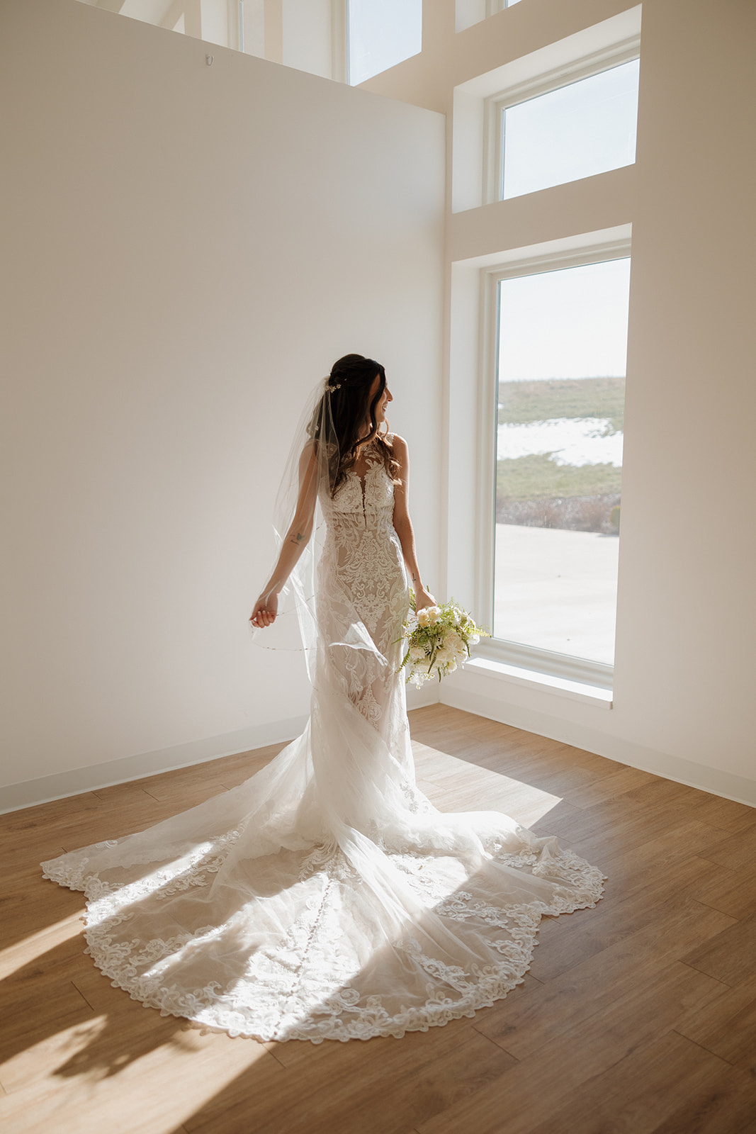 Bride stands alone in a pool of window light, her train fanned out perfectly behind her for an editorial bridal portrait.