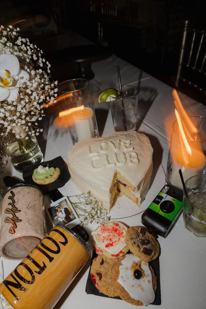 Flat lay of a dessert table with cookies, cocktails, film cameras, and a “Love Club” heart cake.