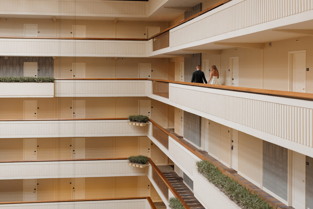 Bride and groom holding hands while walking down a modern hotel hallway.