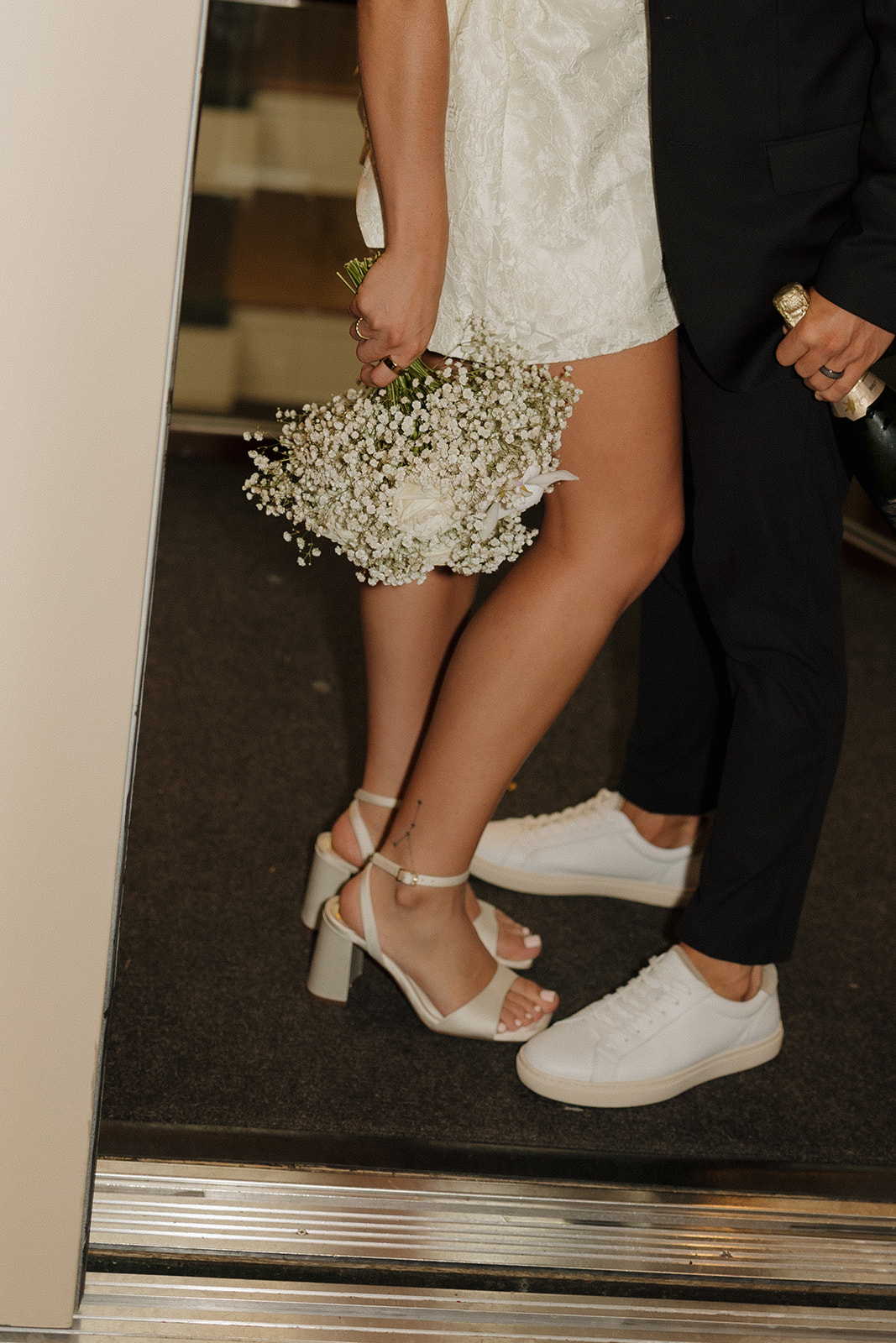 Close-up of a bride and groom’s legs as they stand in an elevator, holding a bouquet of baby’s breath during their wedding in Wisconsin.
