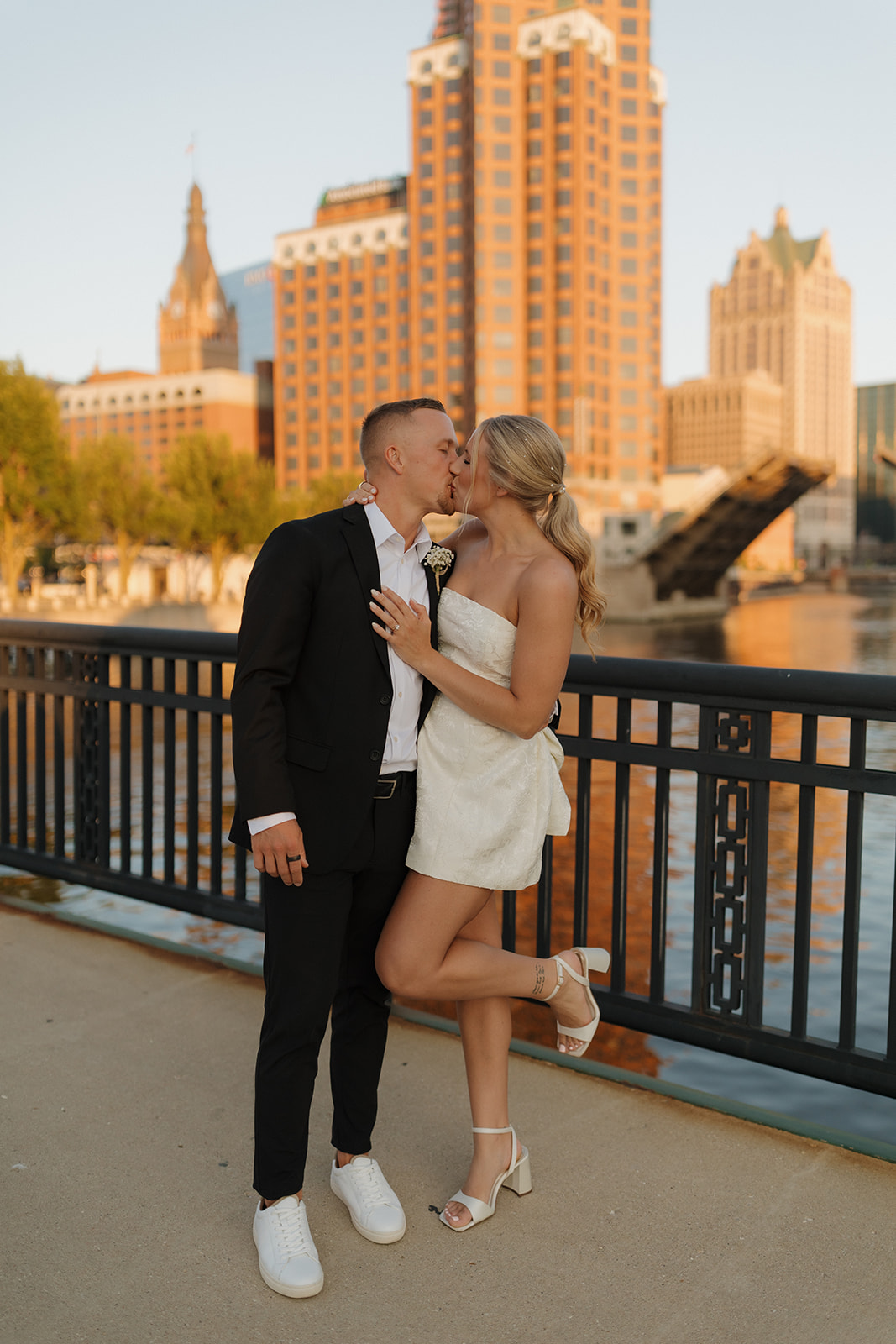 Bride and groom sharing a kiss by the river with Milwaukee’s skyline glowing behind them.