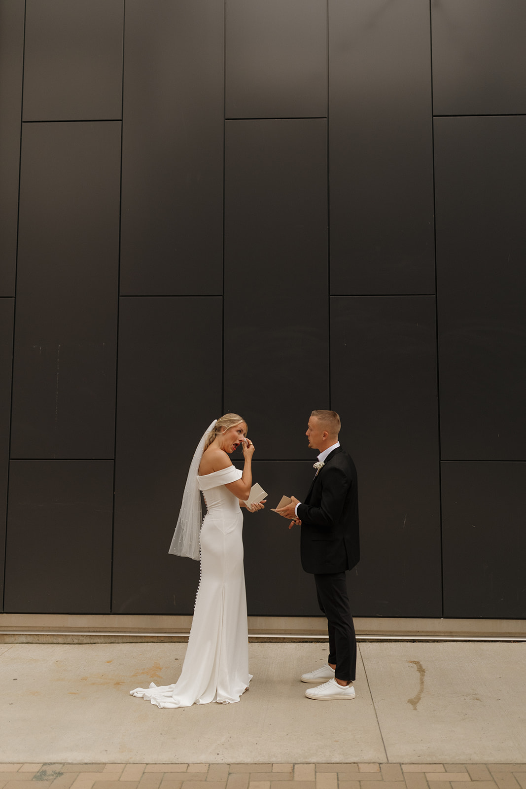Bride wipes tears as groom reads his vows to her during private vow reading in front of black wall in downtown Wisconsin.