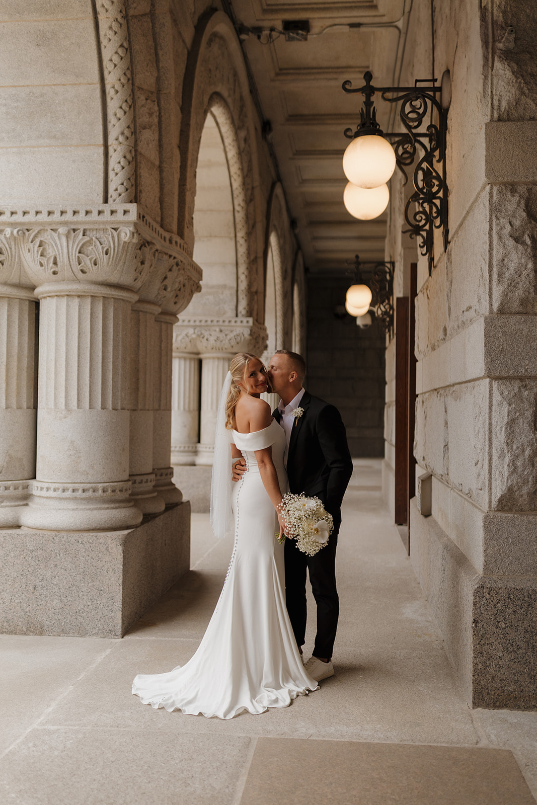 Groom kisses his bride on cheek in secluded marble corner during outdoor bridal portraits