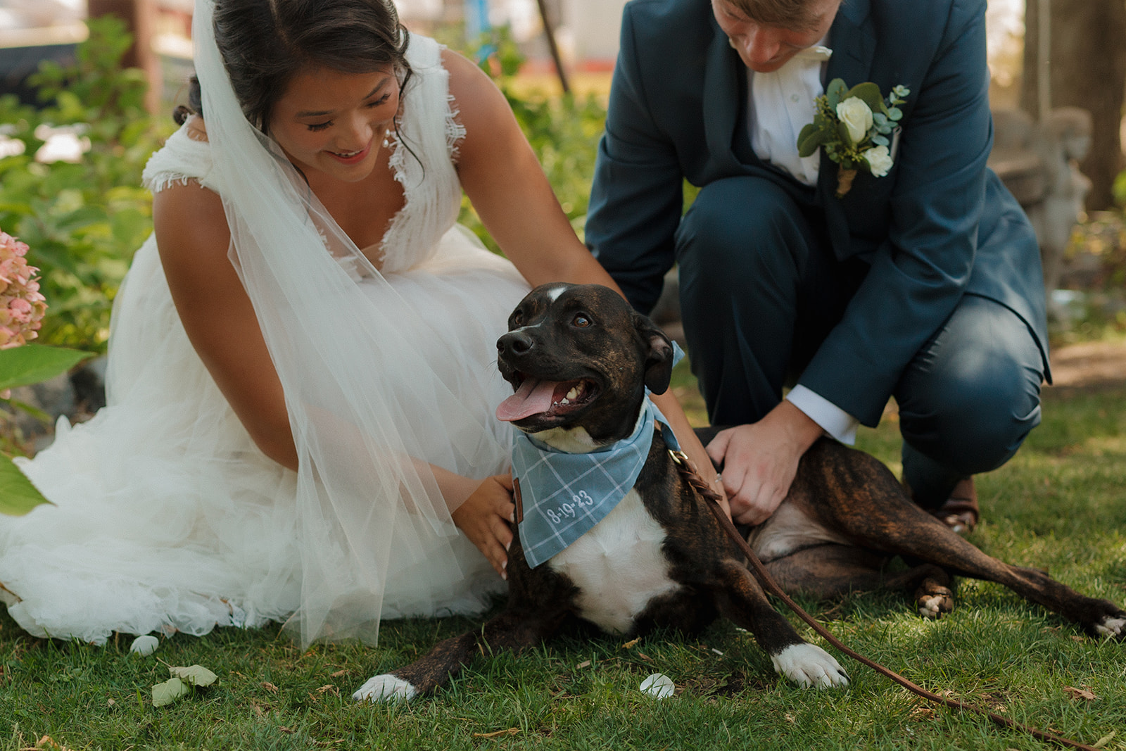 Bride and groom crouched in the grass, laughing with their joyful pup wearing a custom bandana.