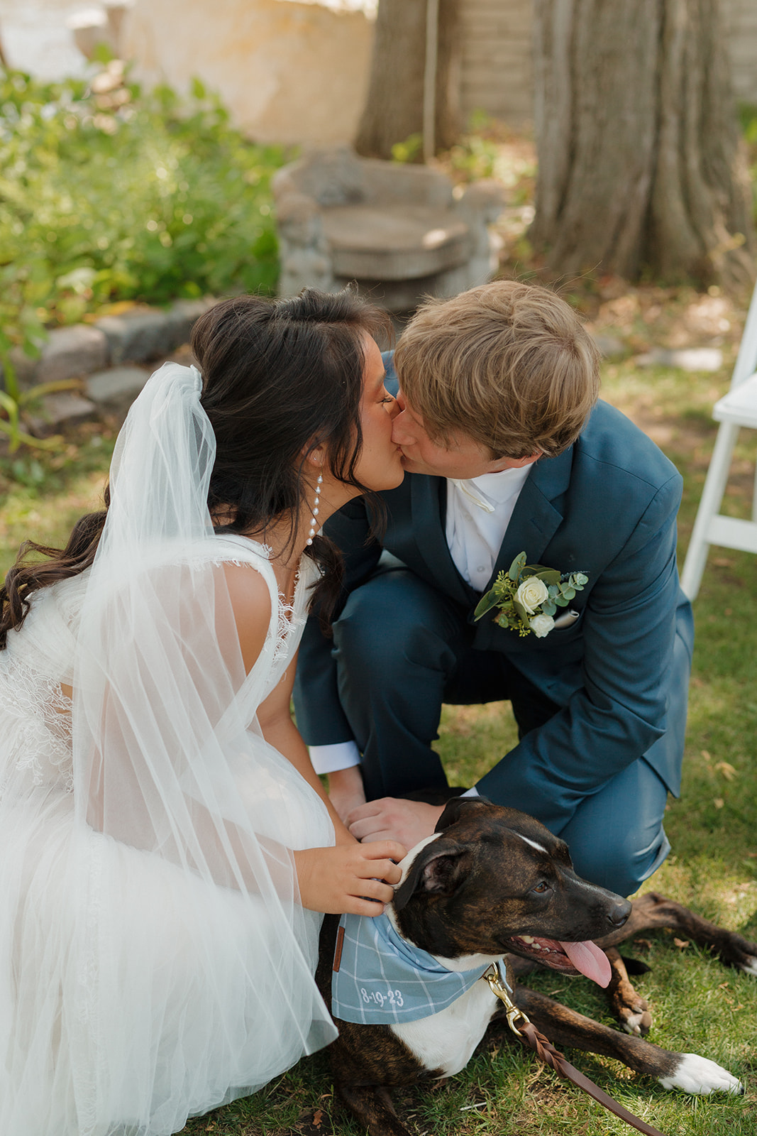 Couple shares a kiss while crouched beside their dog during a candid moment at their outdoor Milwaukee wedding venue.