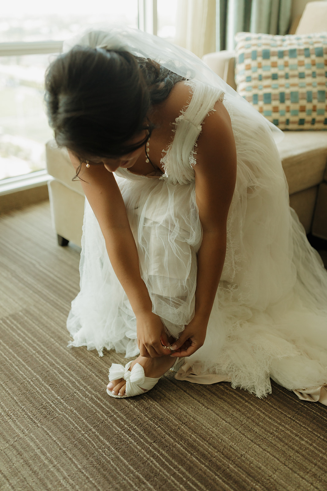 Bride wearing wedding gown and veil bends over to buckle wedding shoes before wedding ceremony