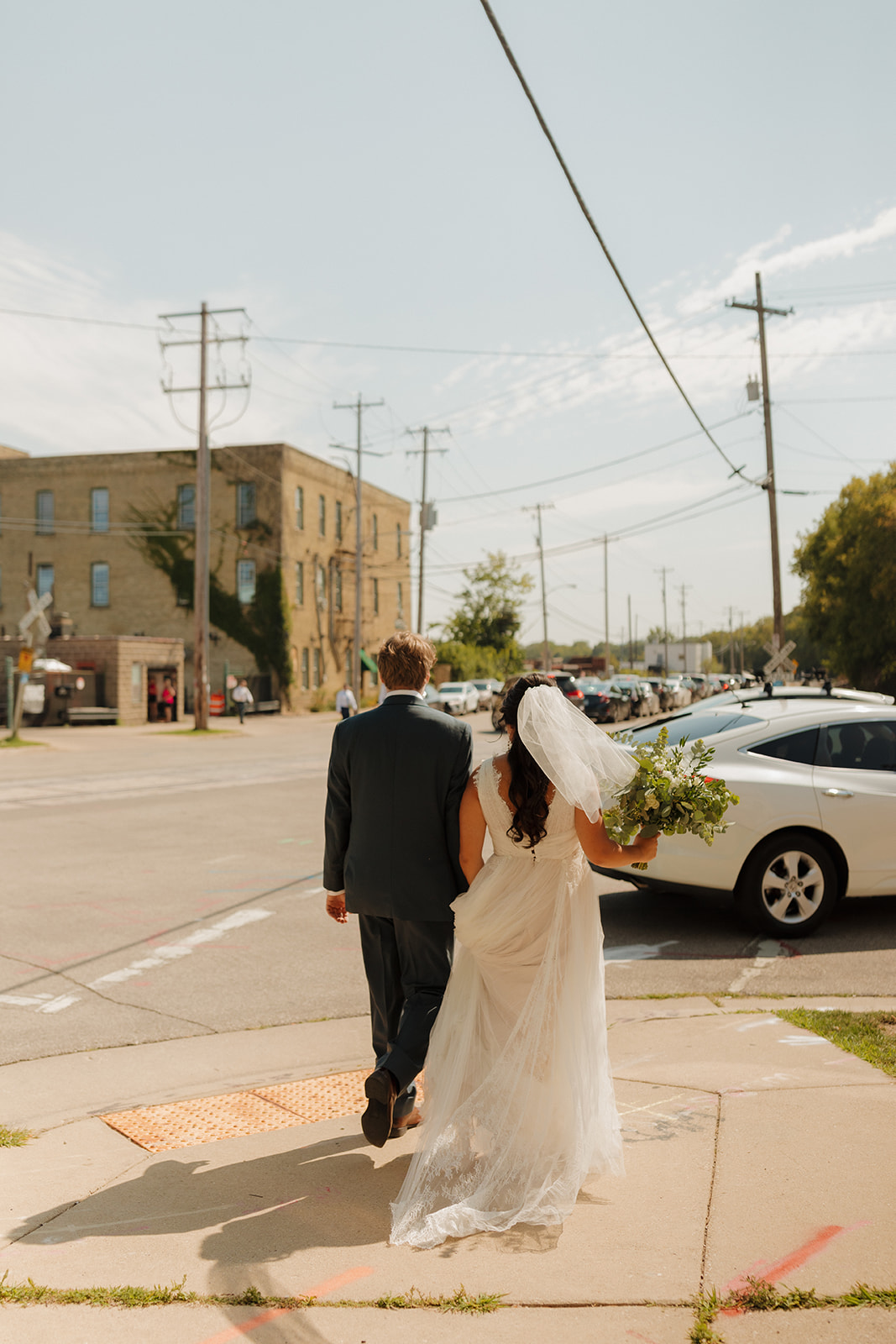 Bride and groom walking hand in hand through an industrial street in Milwaukee, her dress trailing behind and bouquet in hand.