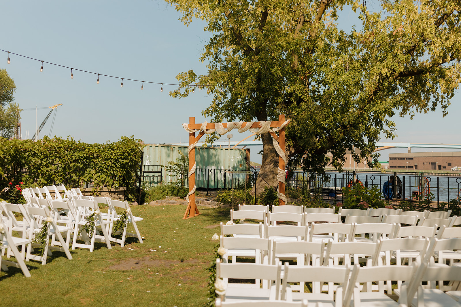 Outdoor ceremony setup at a Milwaukee wedding venue, with white chairs, string lights, and a wooden arch facing the water.