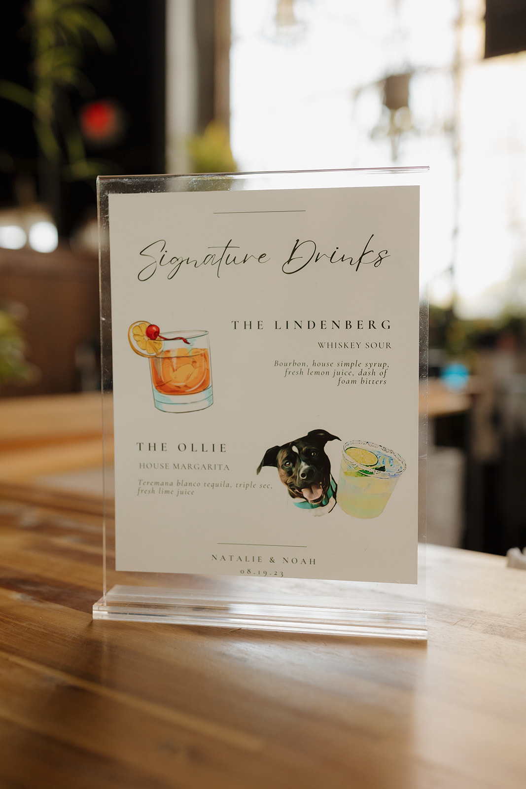 Signature drinks sign on a wooden bar top featuring watercolor illustrations of cocktails and a dog, photographed inside a Milwaukee wedding venue.