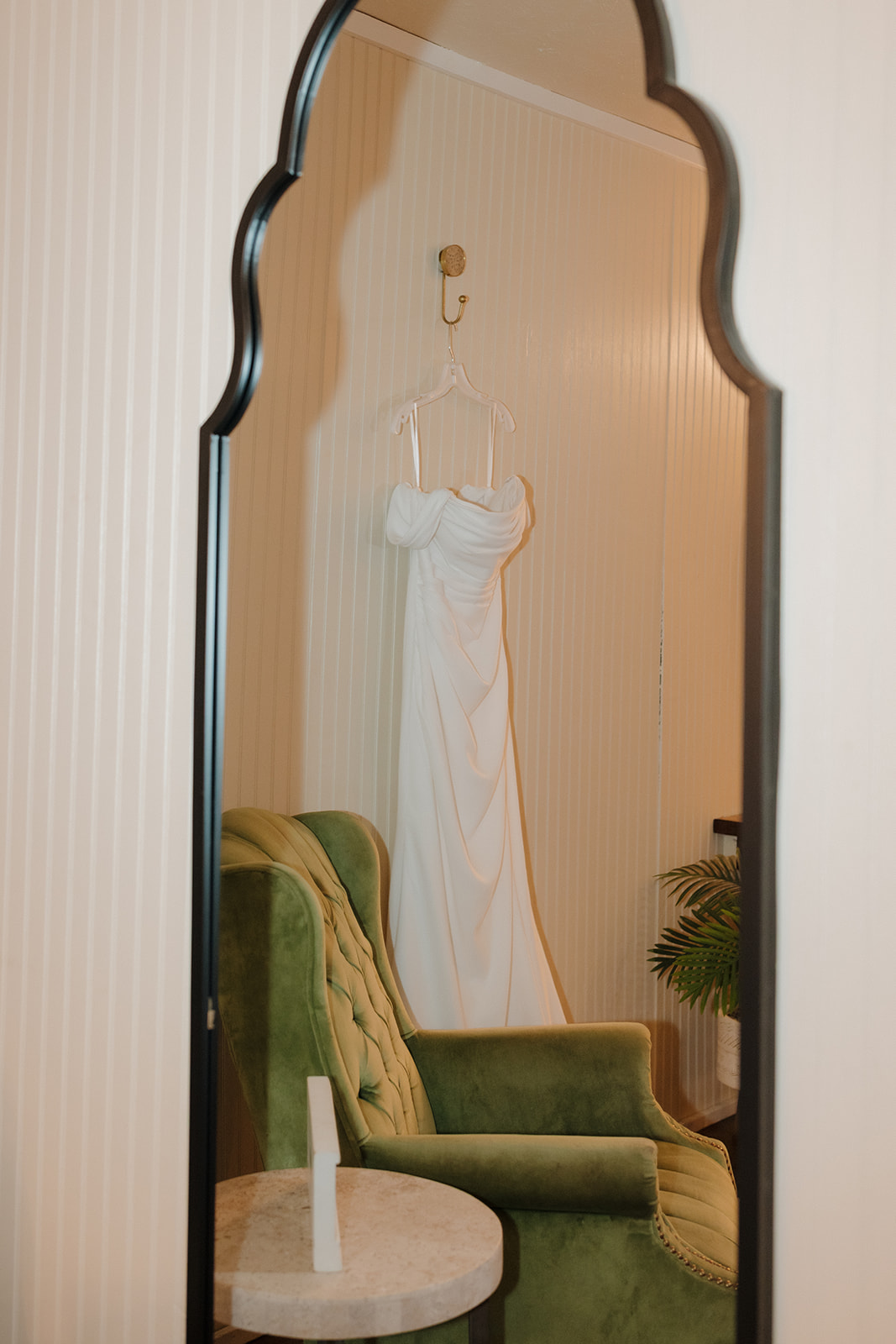 Classic white wedding gown reflected in a tall mirror with a green vintage chair nearby.