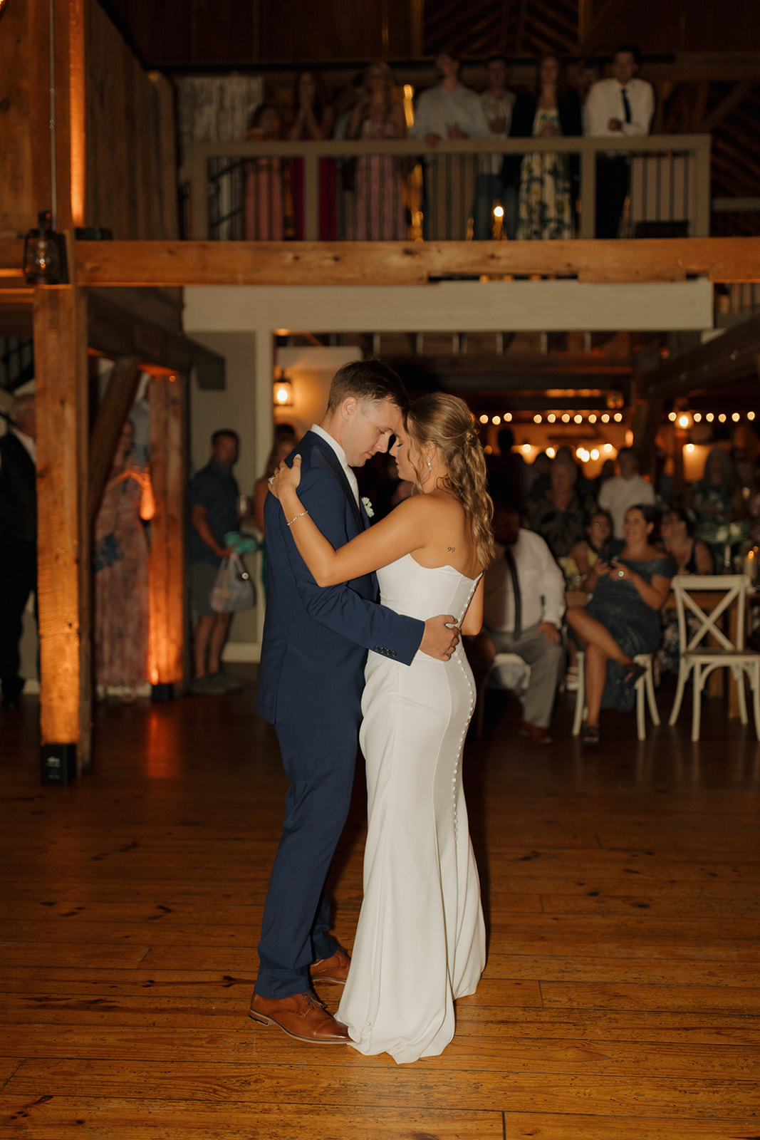 Bride and groom sharing their first dance as guests look on from the candlelit barn reception space.