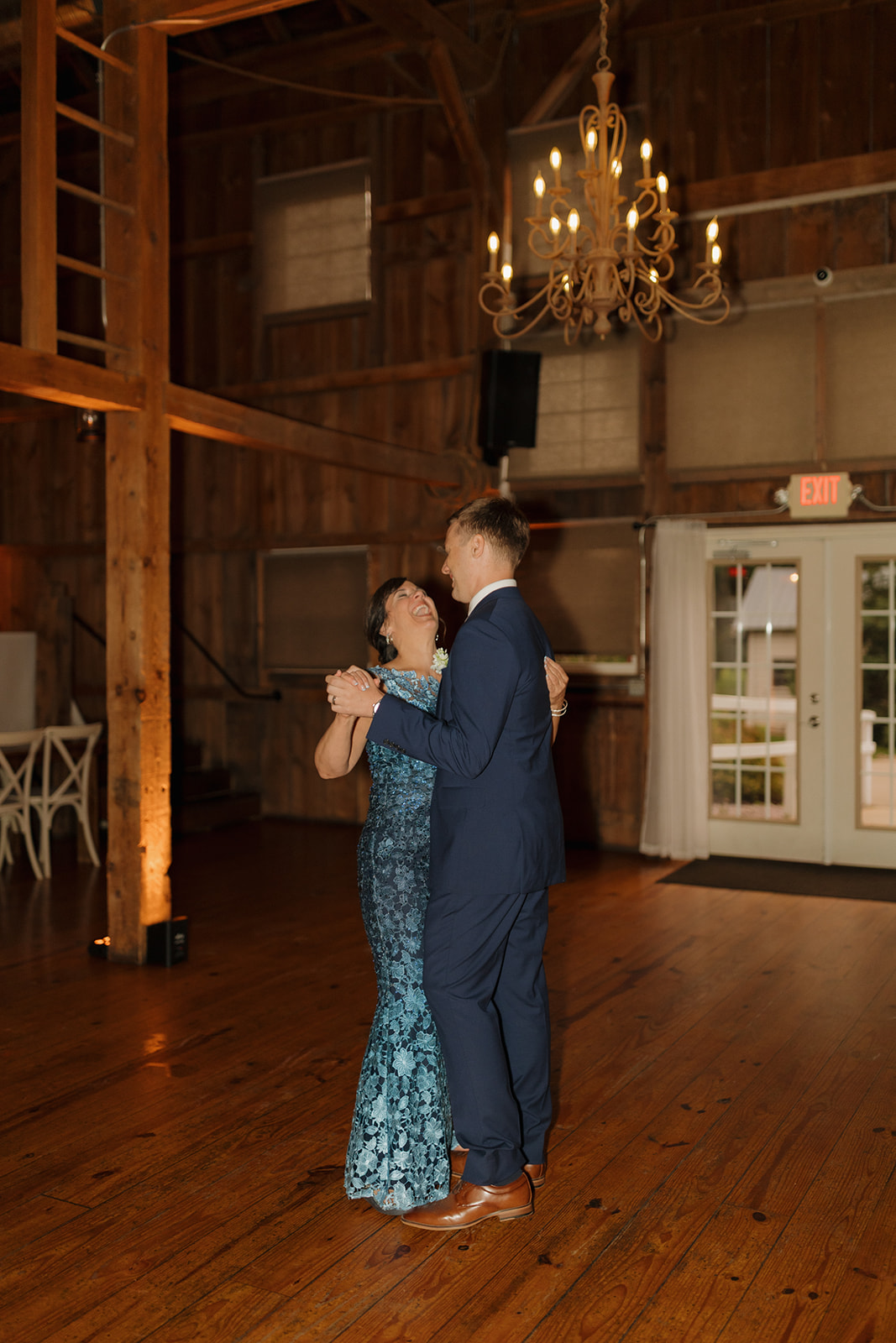 Groom dancing with his mom on the wooden floor of a warmly lit barn venue.