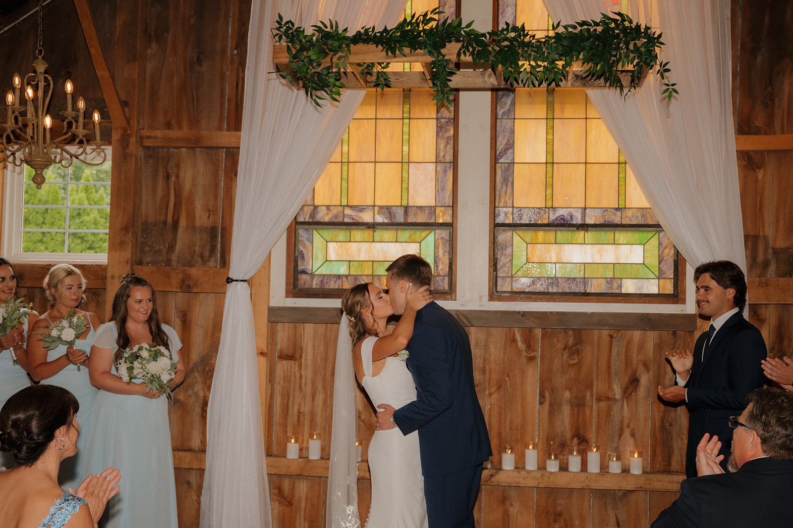 First kiss in front of stained glass windows surrounded by bridesmaids and candles at a wood-paneled ceremony space.