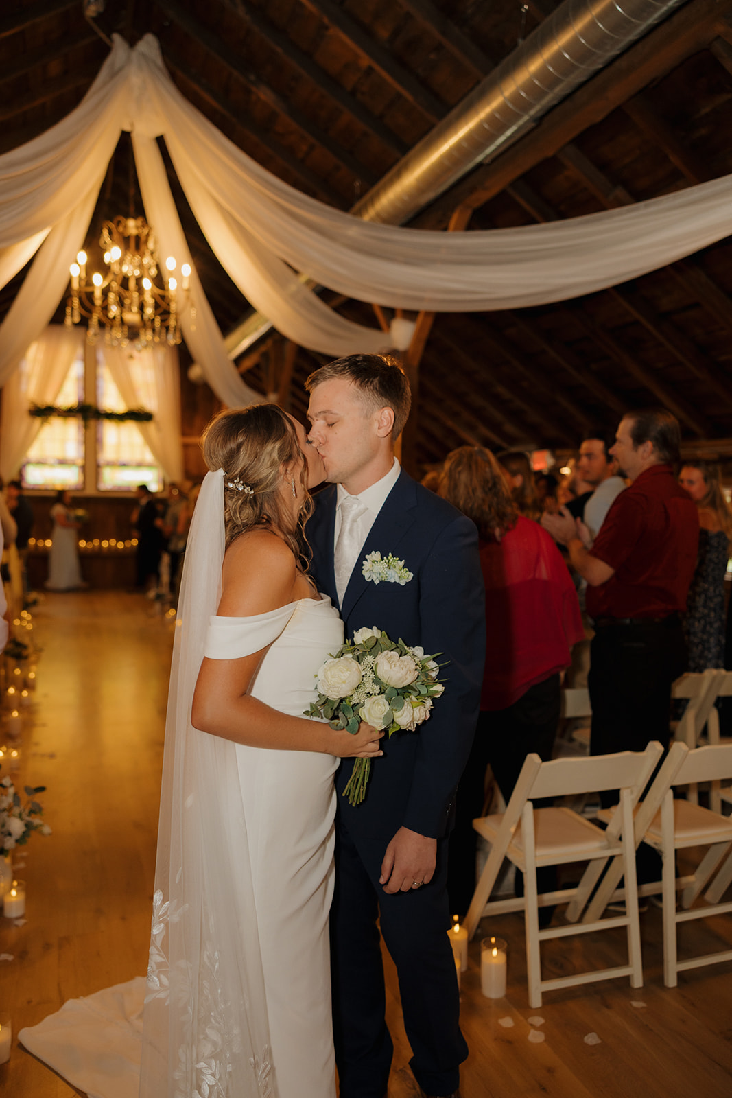Bride and groom kiss at the end of the aisle beneath flowing white drapes and a chandelier.