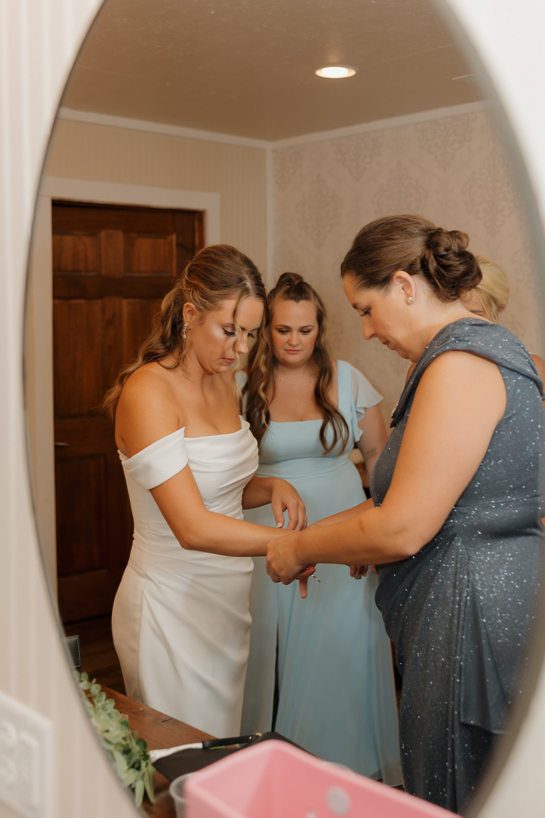 Bride getting ready with the help of her mom and bridesmaids in a soft-lit prep room.