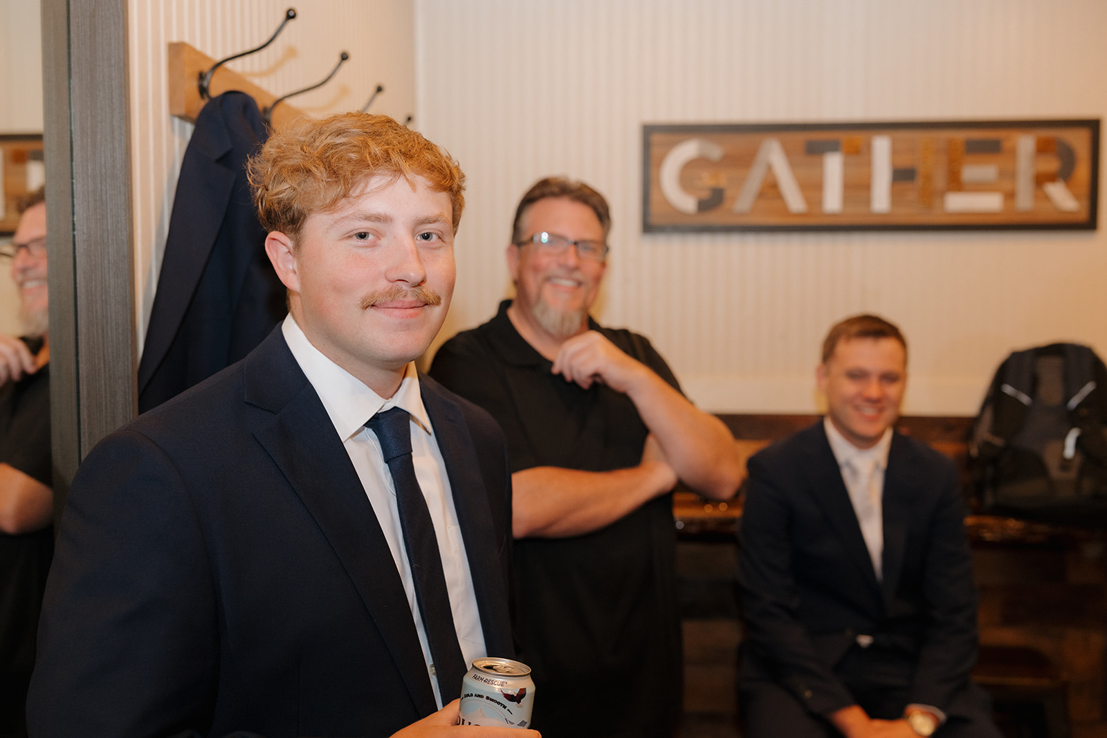 Groomsman smiling while holding a drink, with the groom and friends laughing in the background.