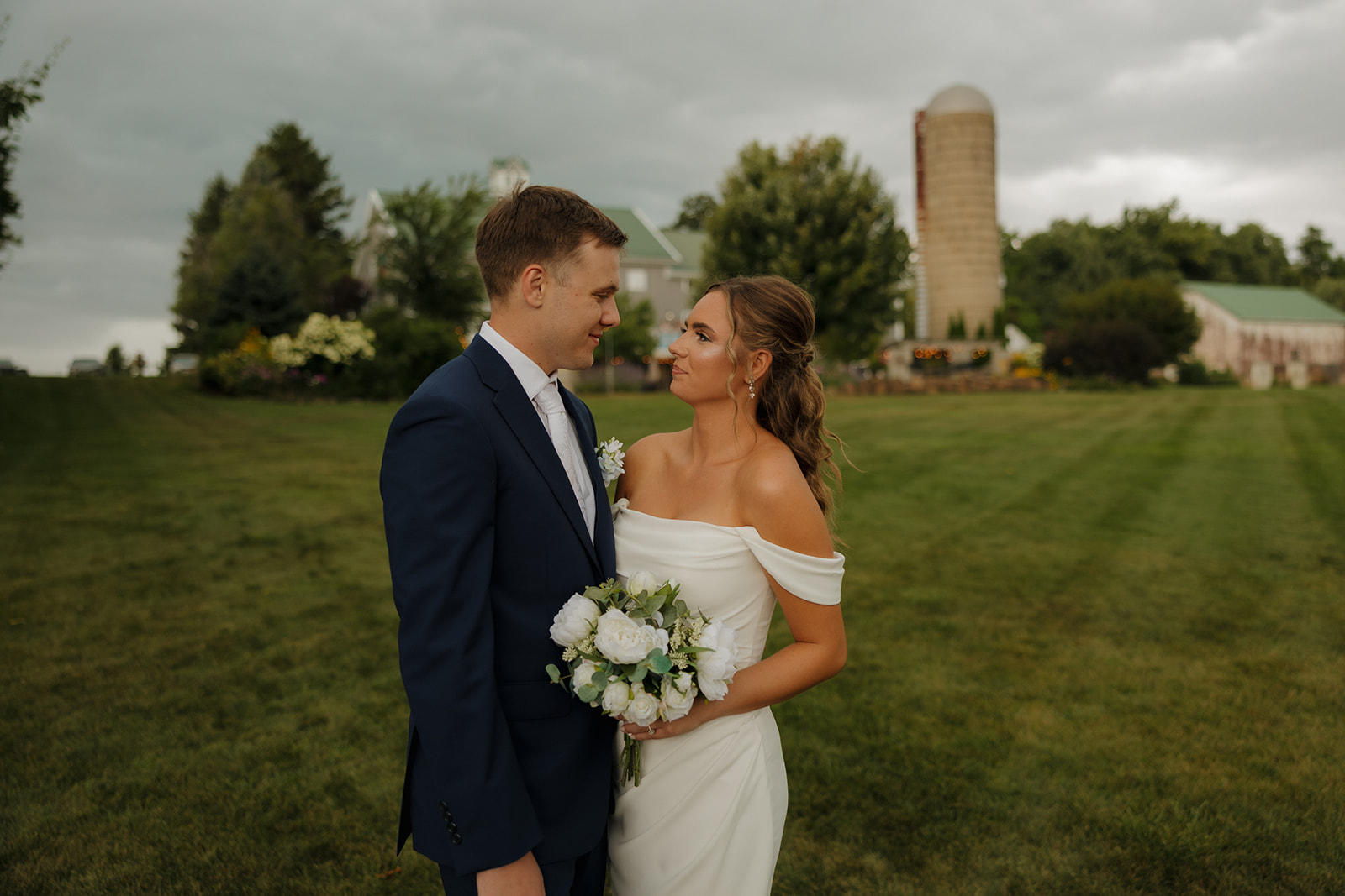 Bride and groom sharing a quiet moment on a wide lawn with a silo and barn in the background.